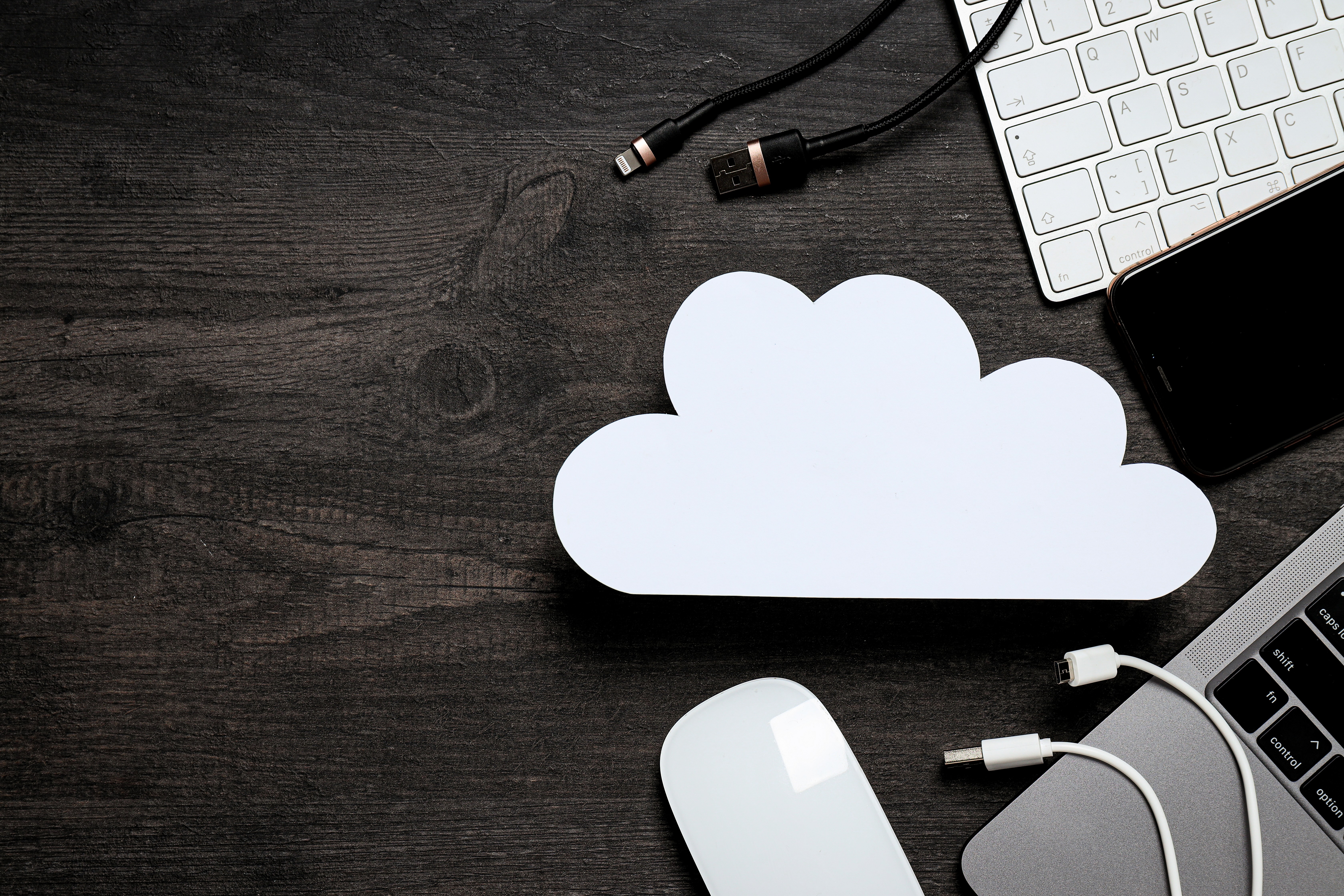 Aus der Vogelperspektive ist eine weiße, aus Papier geschnittene Wolke auf einem dunklen Holztisch zu sehen. Um das Cloud-Symbol herum sind verschiedene technische Geräte wie ein Laptop, ein Smartphone, eine Tastatur, eine Maus und Ladekabel angeordnet, die das Konzept des Cloud-Computing symbolisieren.