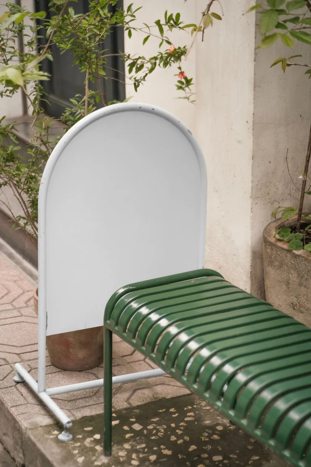 Mockup of a freestanding signage board placed next to a green metal bench in a quaint outdoor setting. Surrounded by potted plants and natural elements, it offers a cozy and inviting aesthetic for branding presentations.