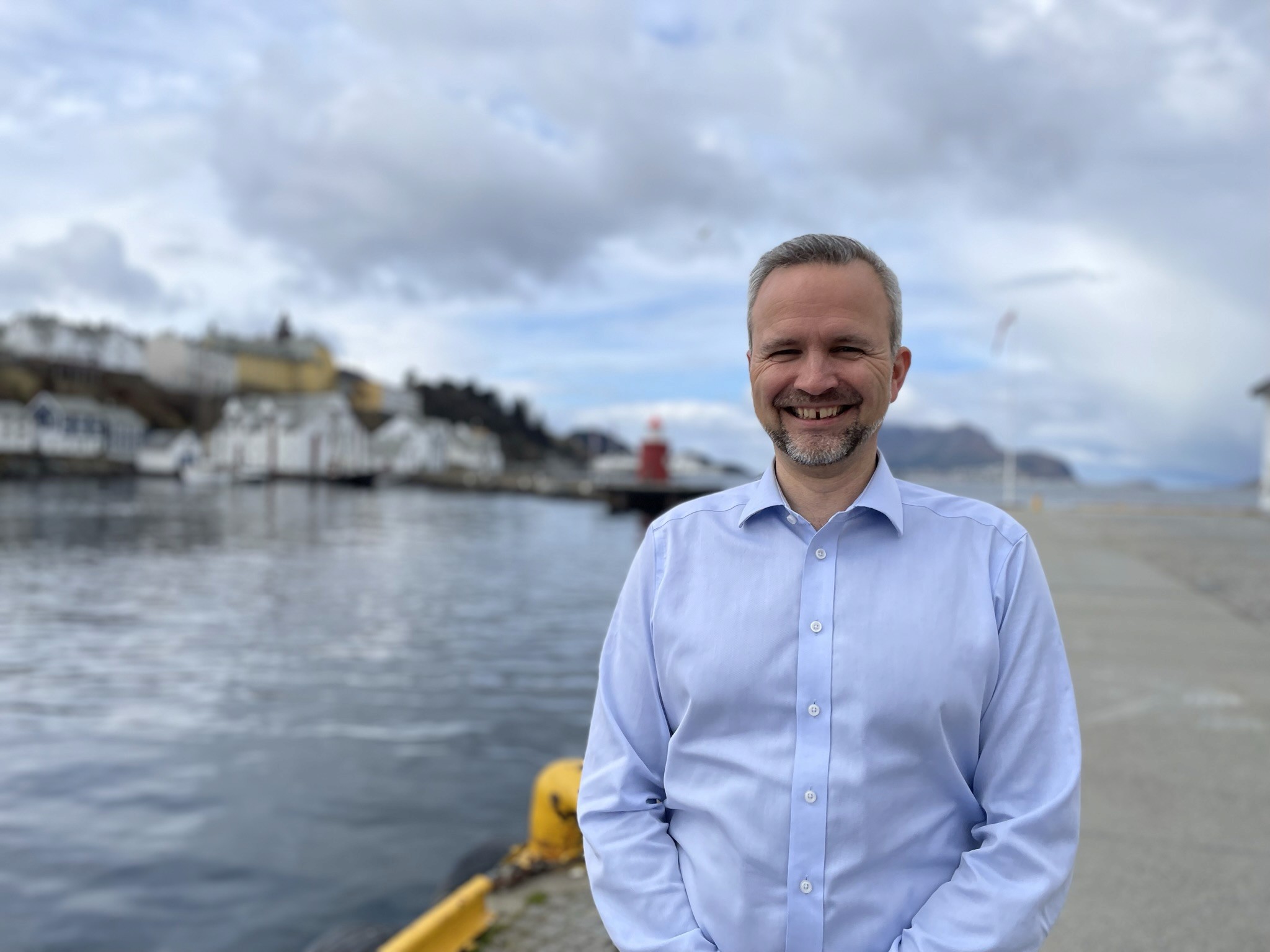A smiling man stands by a waterfront, wearing a light blue shirt, with buildings and cloudy sky in the background.