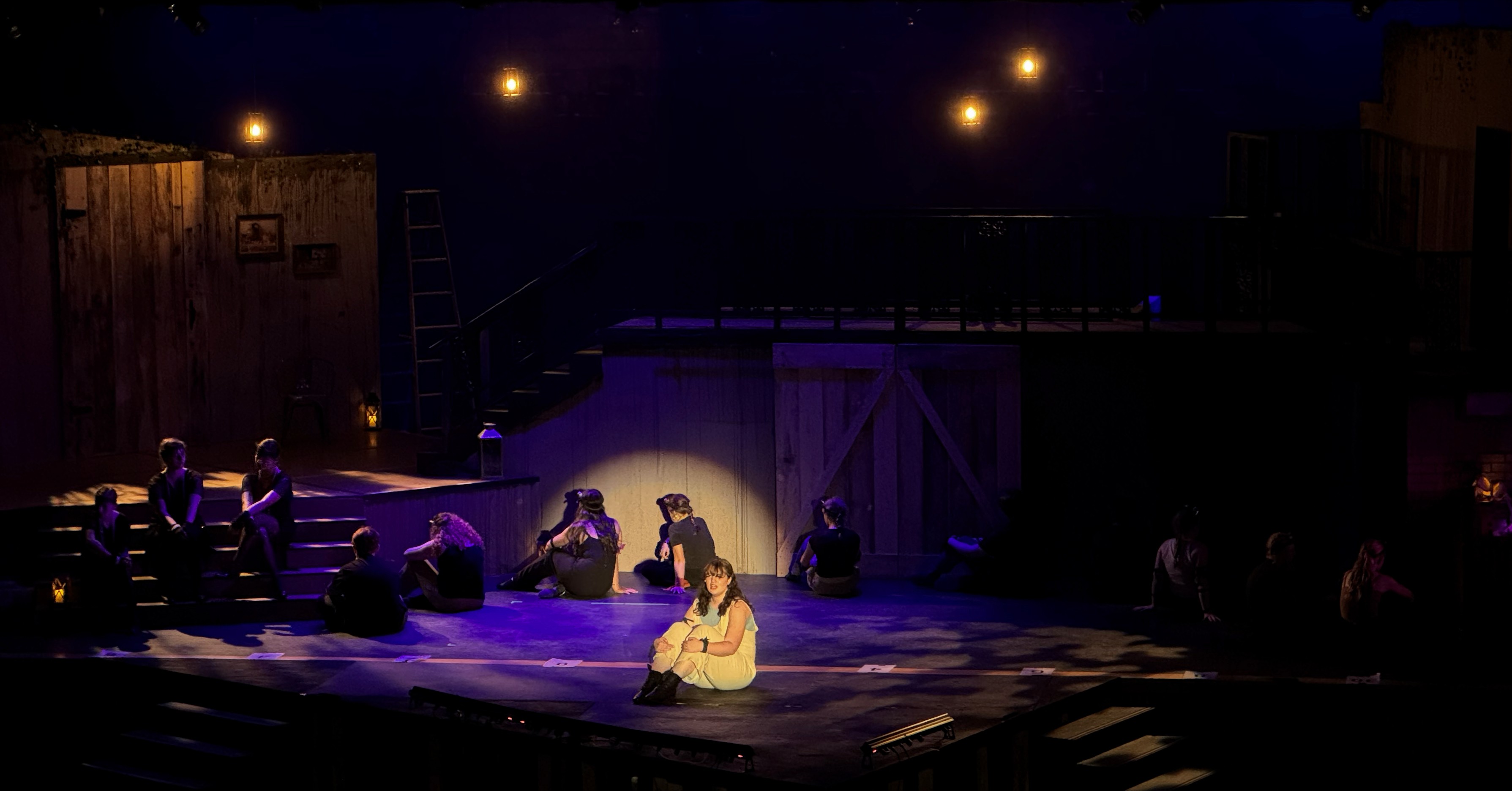 Eurydice sits alone in a pool of blue light, surrounded by hanging laterns and intricate breakup gobos patterns, creating an intimate spotlight effect on a dimly lit stage with other cast members seated in the shadows.