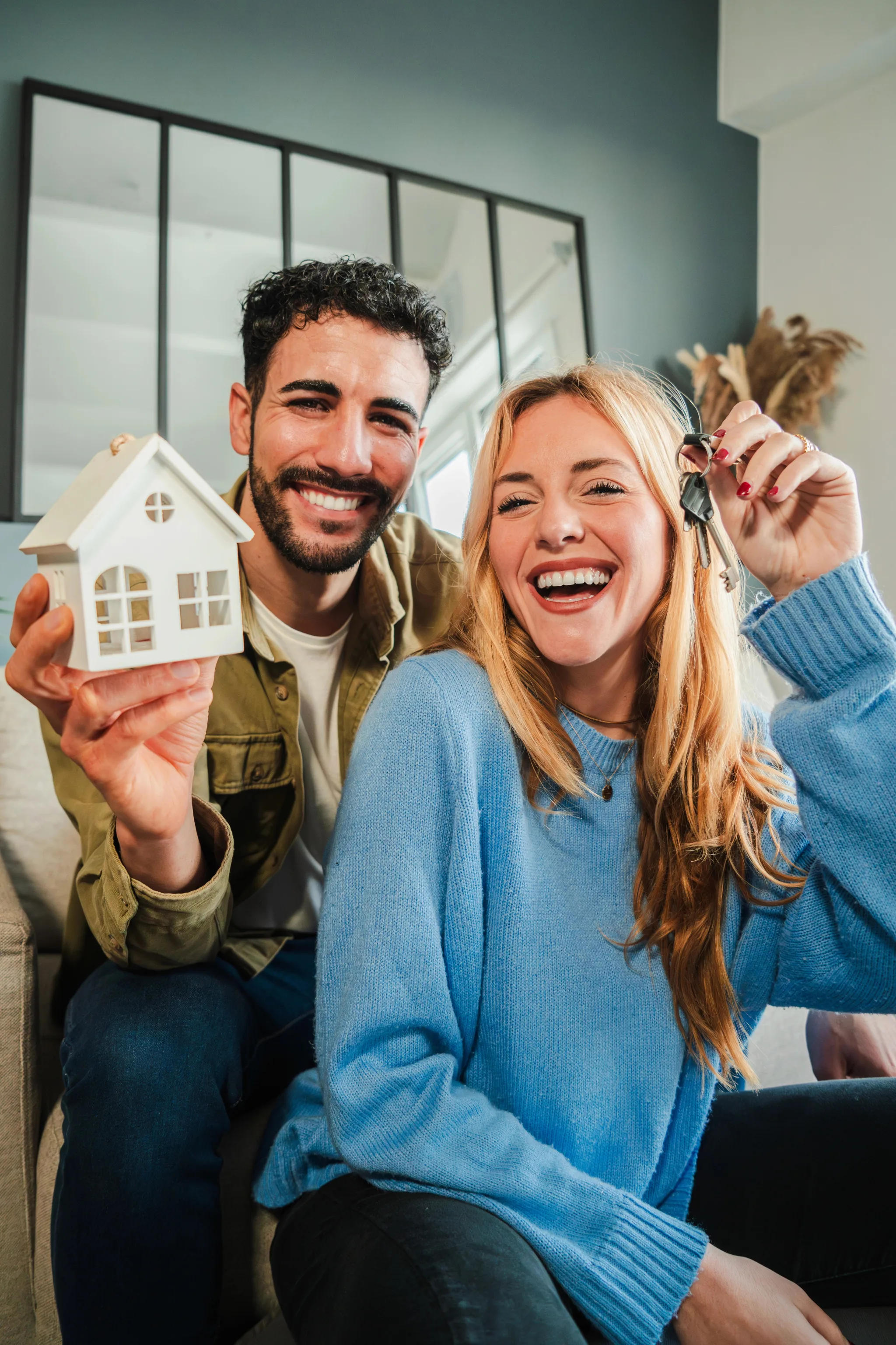 A smiling young couple celebrates holding a miniature white house model and keys in their modern home with sage green walls and industrial windows.