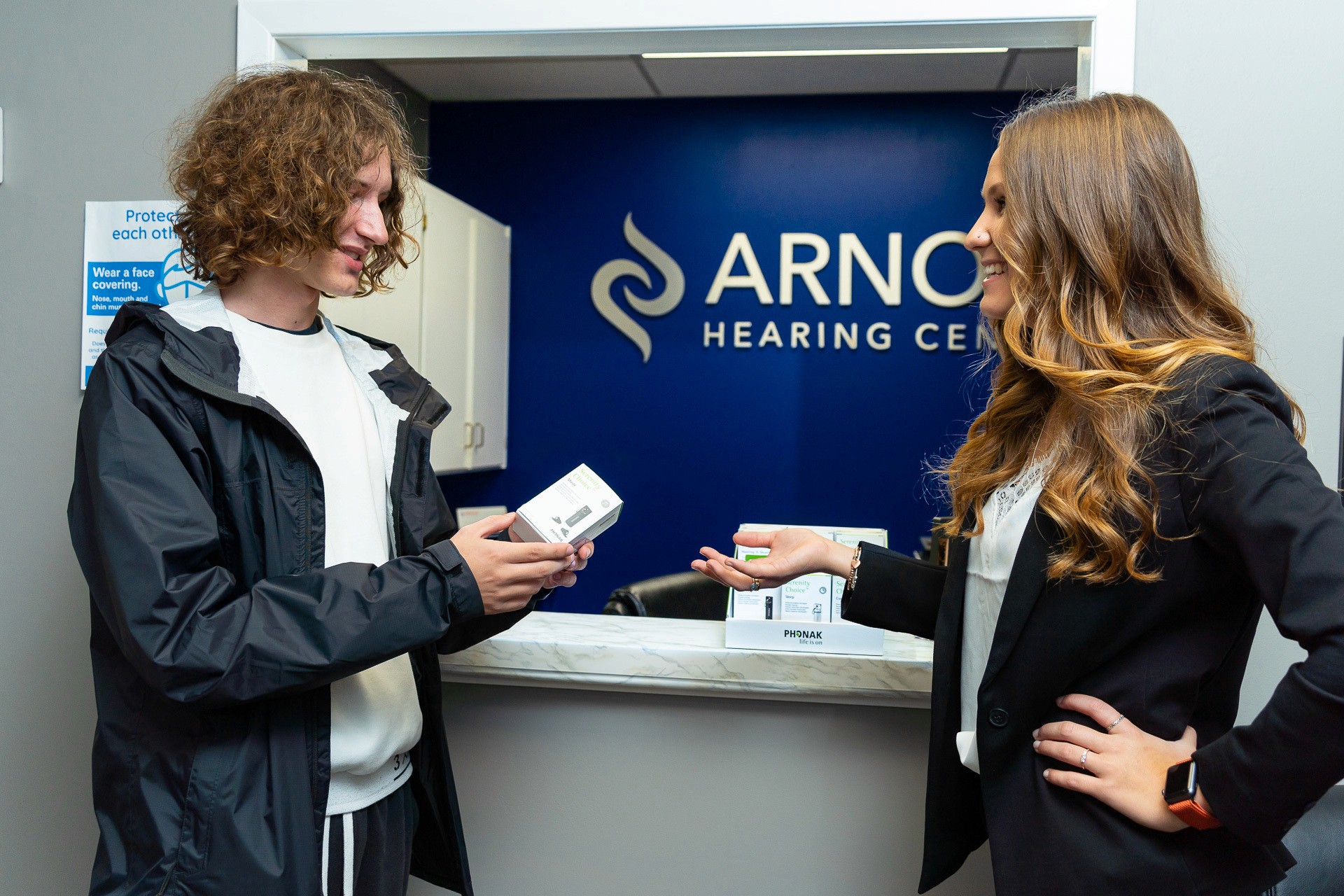 A man and woman stand at a service counter, discussing something while facing a blue company sign behind them.