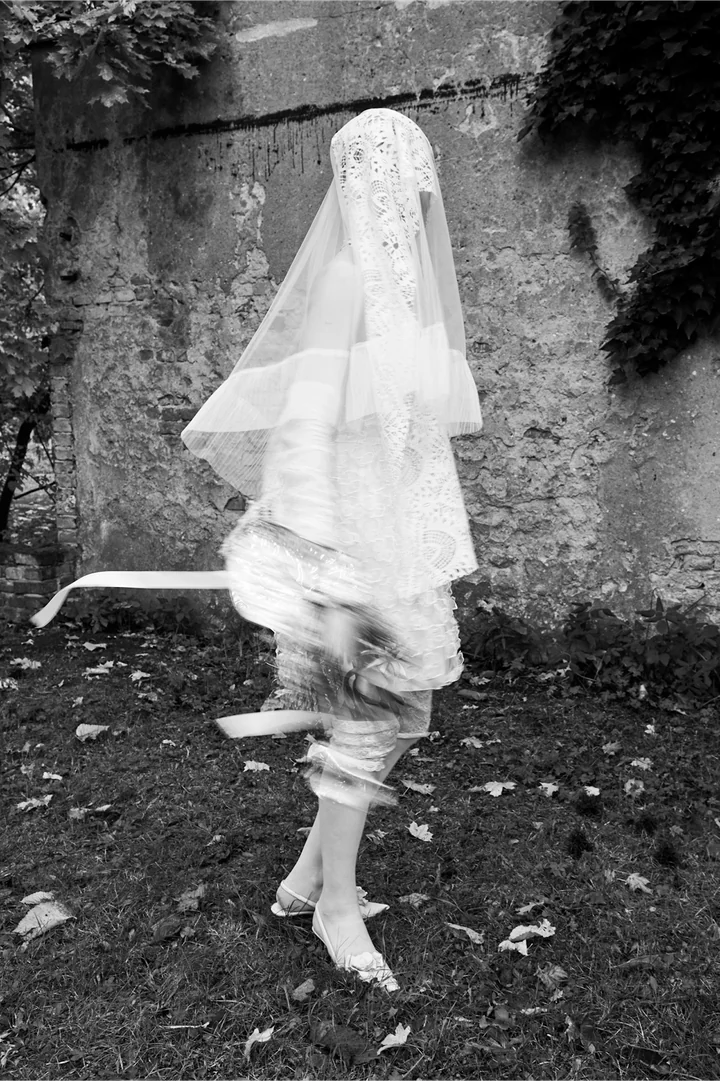 Black and white photo of a bride in a veil twirling barefoot on leaves in front of a stone wall, her dress blurred with motion.