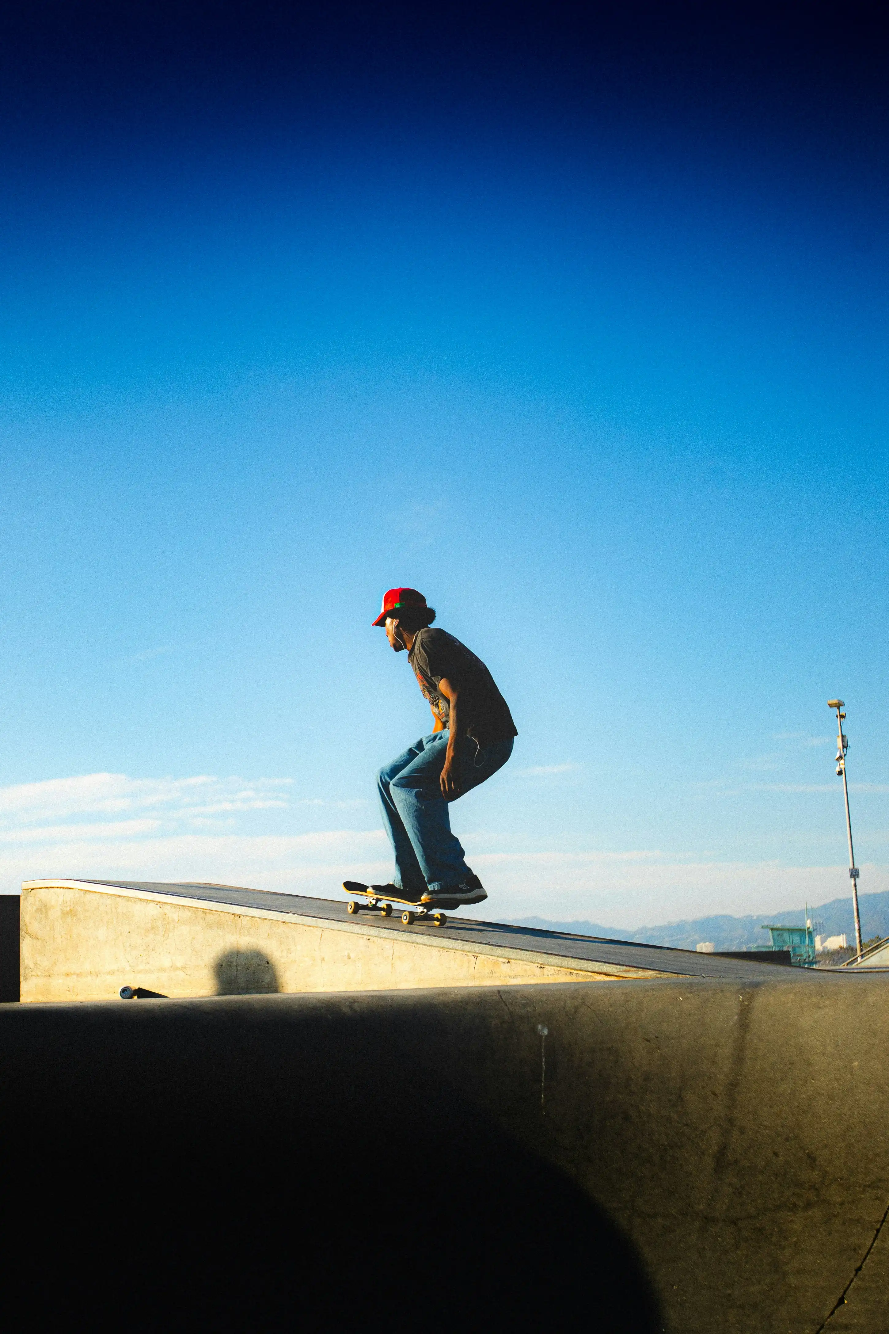A person wearing a red cap performing a trick on a skateboard on a concrete ramp under a bright blue sky.