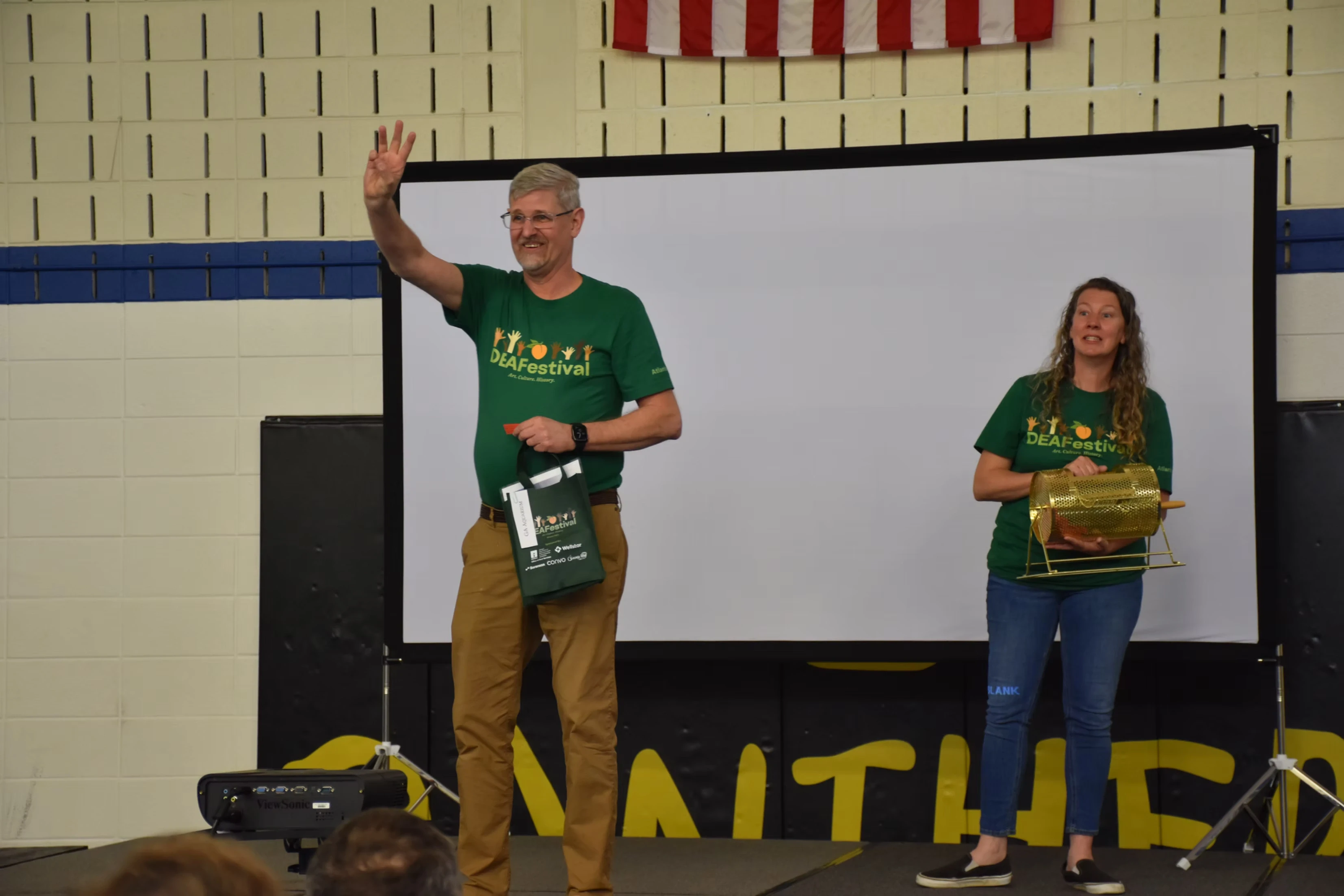 Man in green shirt waves, woman beside him holds a drum. Both are on stage at an event.