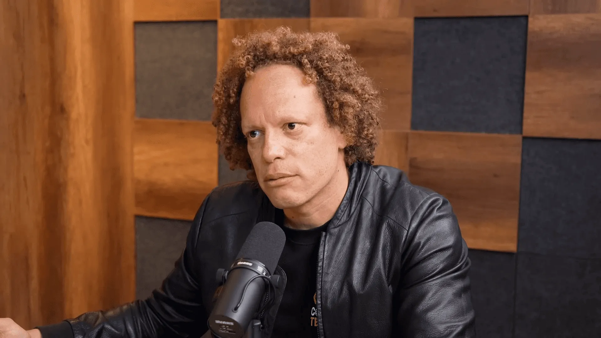 Persona hablando frente a un micrófono en un entorno de grabación o entrevista, con fondo de paneles de madera y vestimenta oscura.
