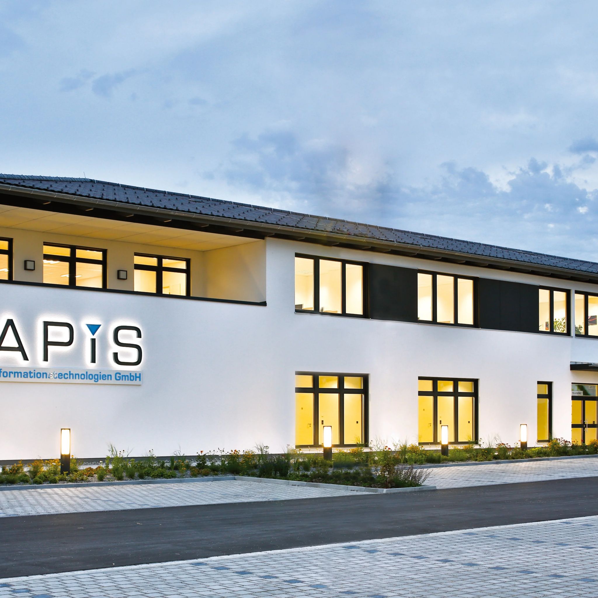 Modernes Bürogebäude mit beleuchtetem APIS Schriftzug, "Informationstechnologien GmbH" Aufschrift und gepflastertemParkplatz.