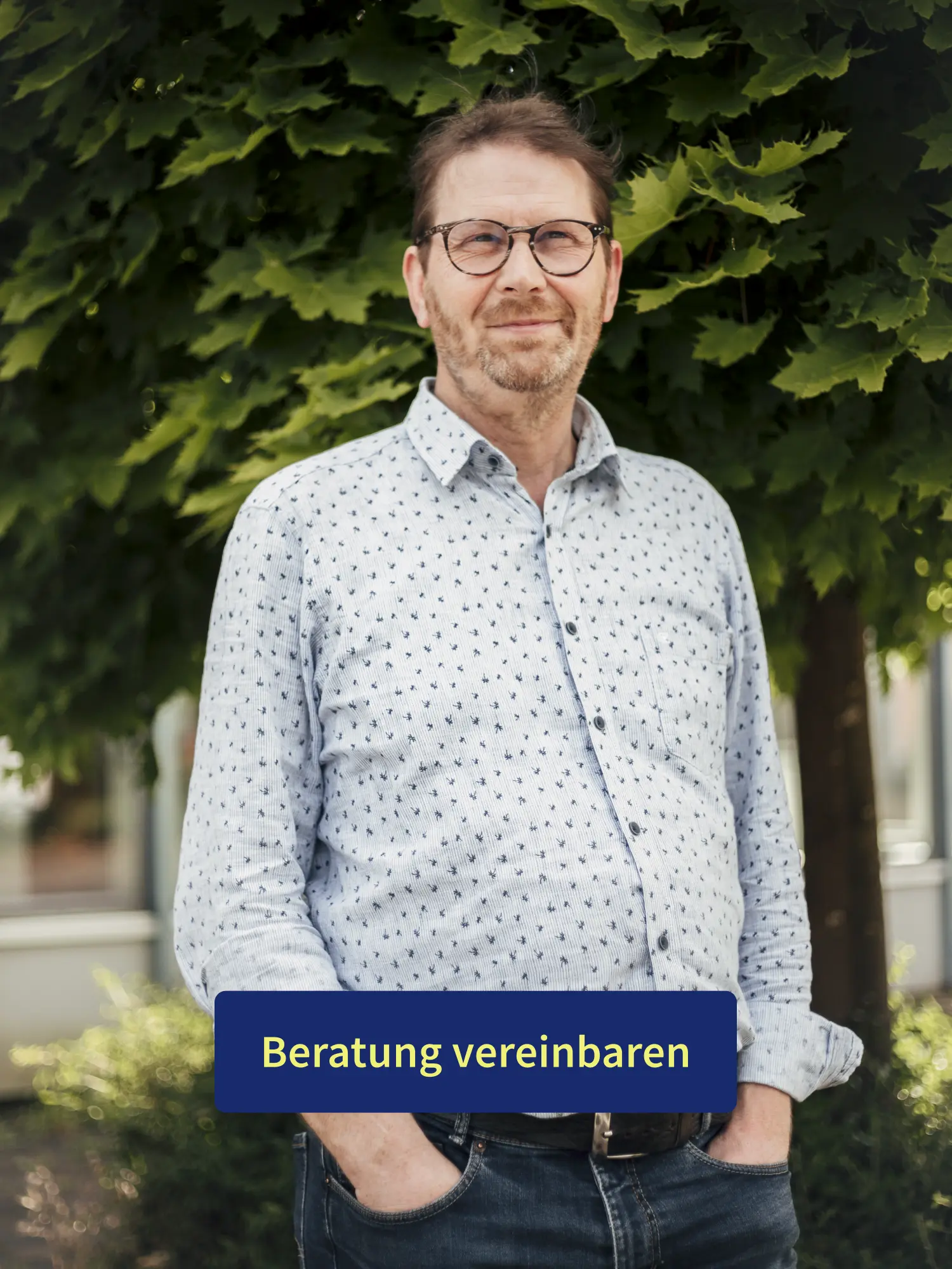 Porträt eines lächelnden Mannes mit Brille vor grünem Baum mit Button „Beratung vereinbaren“.
