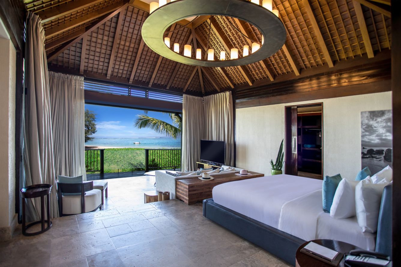 Luxuriöses Schlafzimmer in einer Villa mit hohem Holzdach, einem großen runden Kronleuchter und einer offenen Fensterfront, die den Blick direkt auf den Strand und das Meer freigibt.