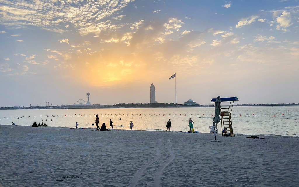 People enjoying a beach at sunset with the Al Forsan Village skyline in the background.