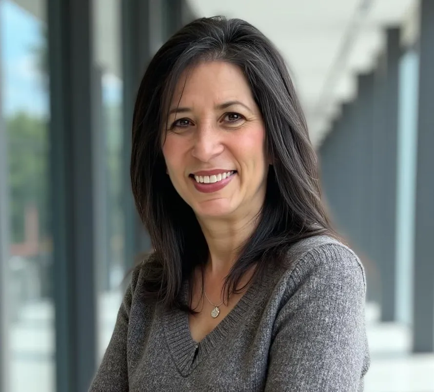 Woman smiling warmly, wearing a gray top, with long dark hair, in a modern indoor setting with large windows.