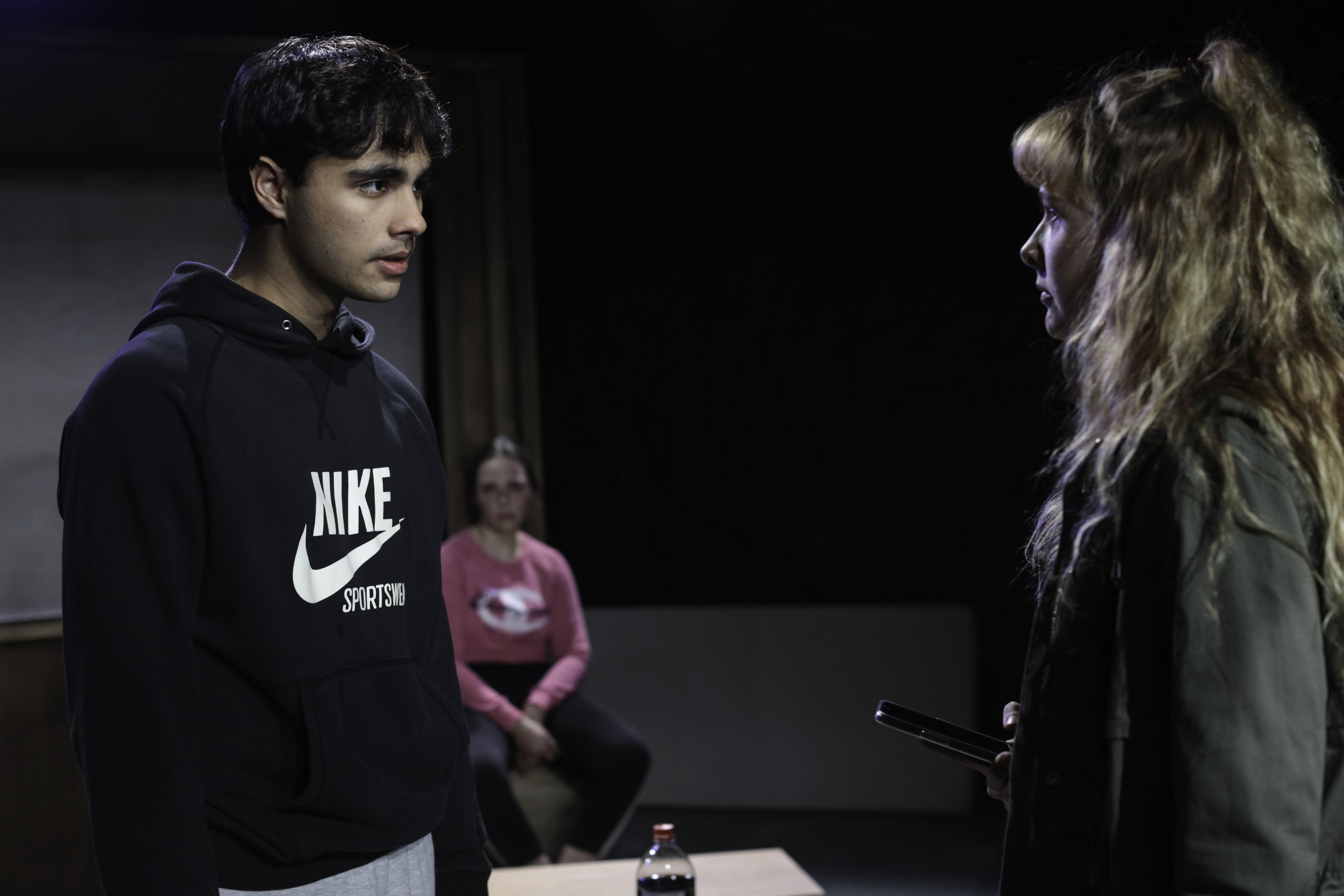 A young man in a dark Nike hoodie faces a young woman holding a phone, their expressions tense. Another woman sits in the background watching them, suggesting a confrontation in a domestic drama setting.
