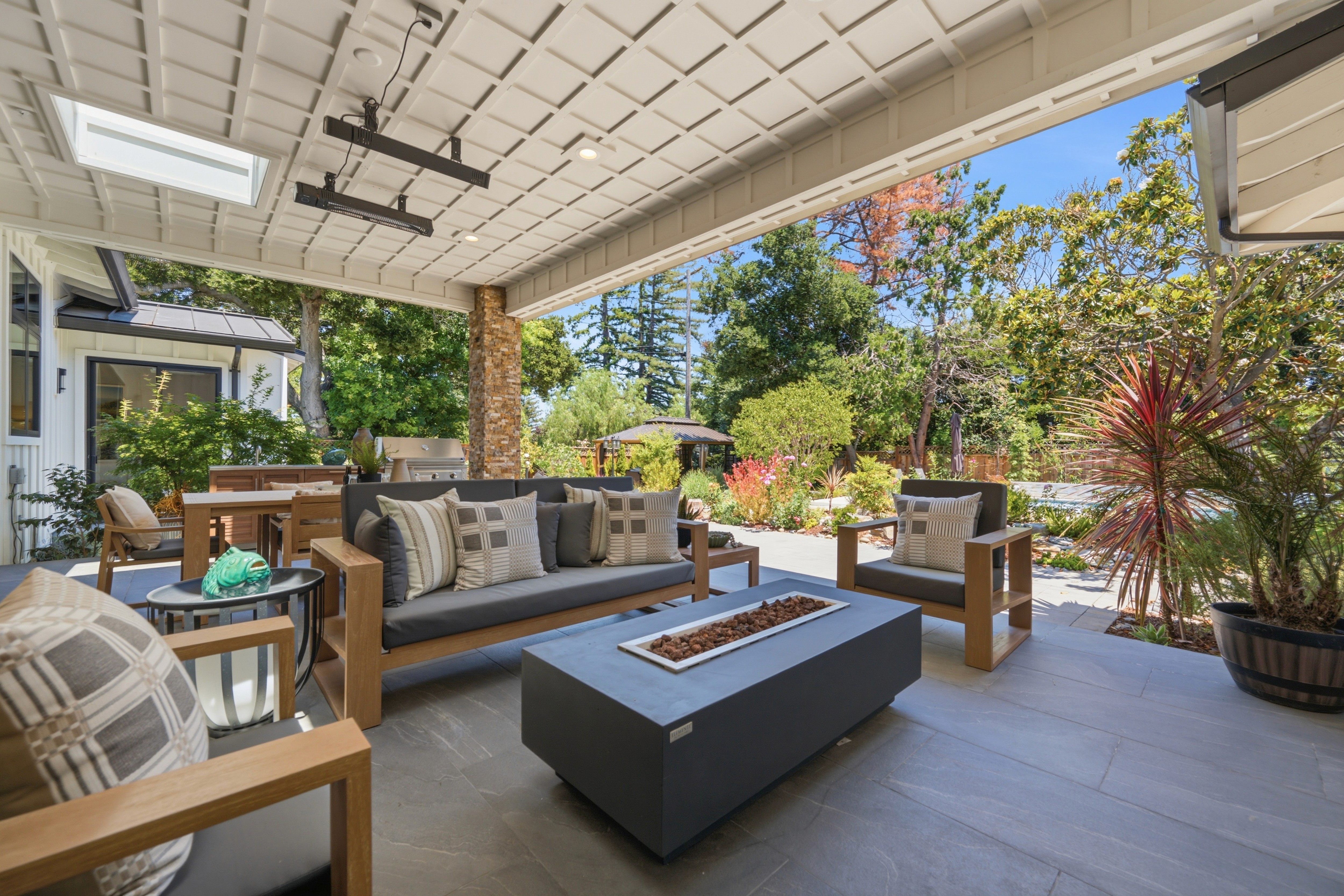 Covered outdoor lounge with a gray fire table, wood-framed sofas, and overhead heaters beneath a coffered ceiling.