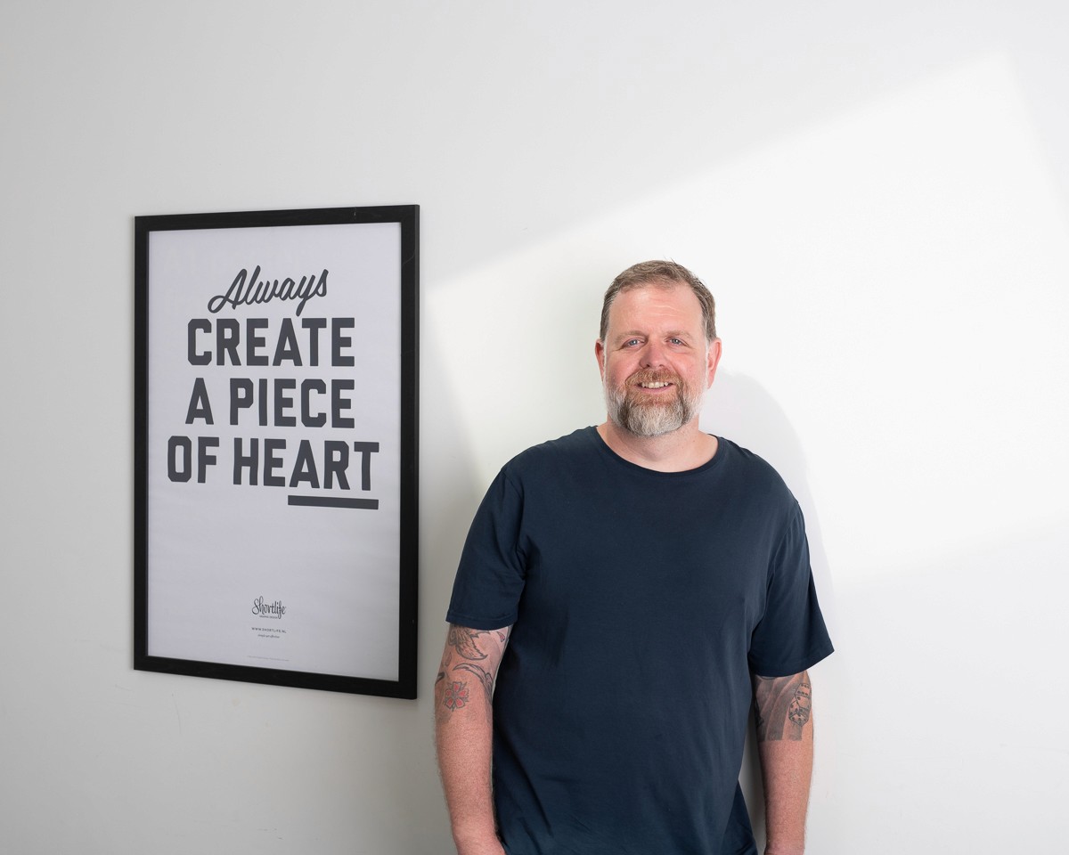 A smiling man with short hair stands with arms crossed, wearing a dark shirt against a neutral background.