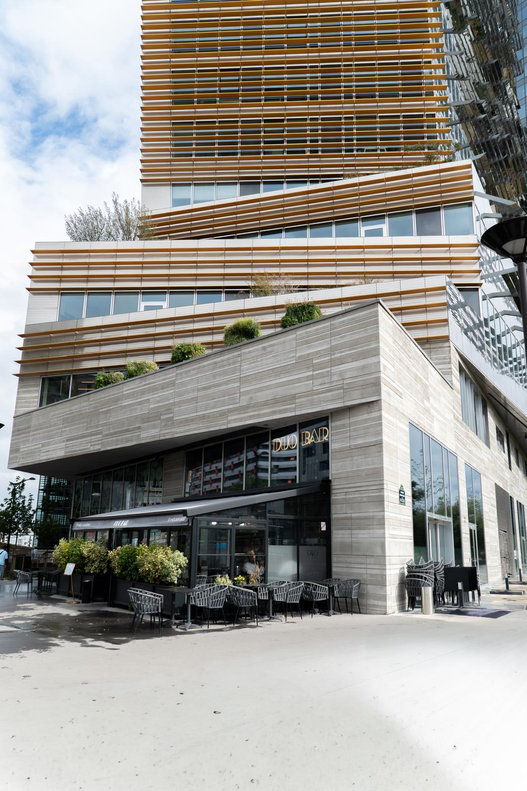 Sunlight streaming through lush green trees, casting shadows on a modern architectural building with glass panels and vertical wooden slats.