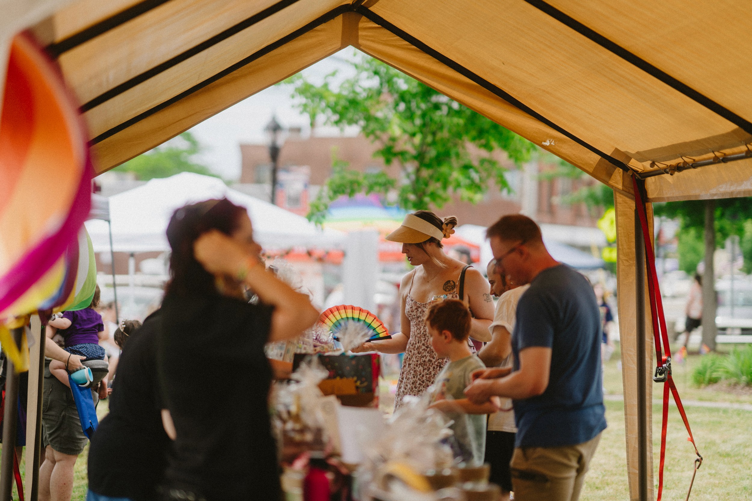 Stoughton Pride vendor tent with attendees gathered around a raffle basket table and the downtown brick architecture in the distance.
