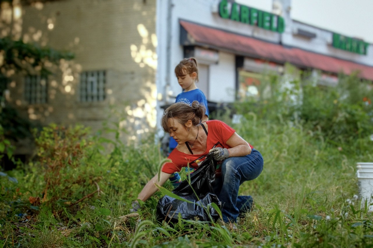 A woman wearing a red shirt and gloves crouches down to gather weeds and debris into a black trash bag, while a child stands nearby, in a lush overgrown area outside a market.