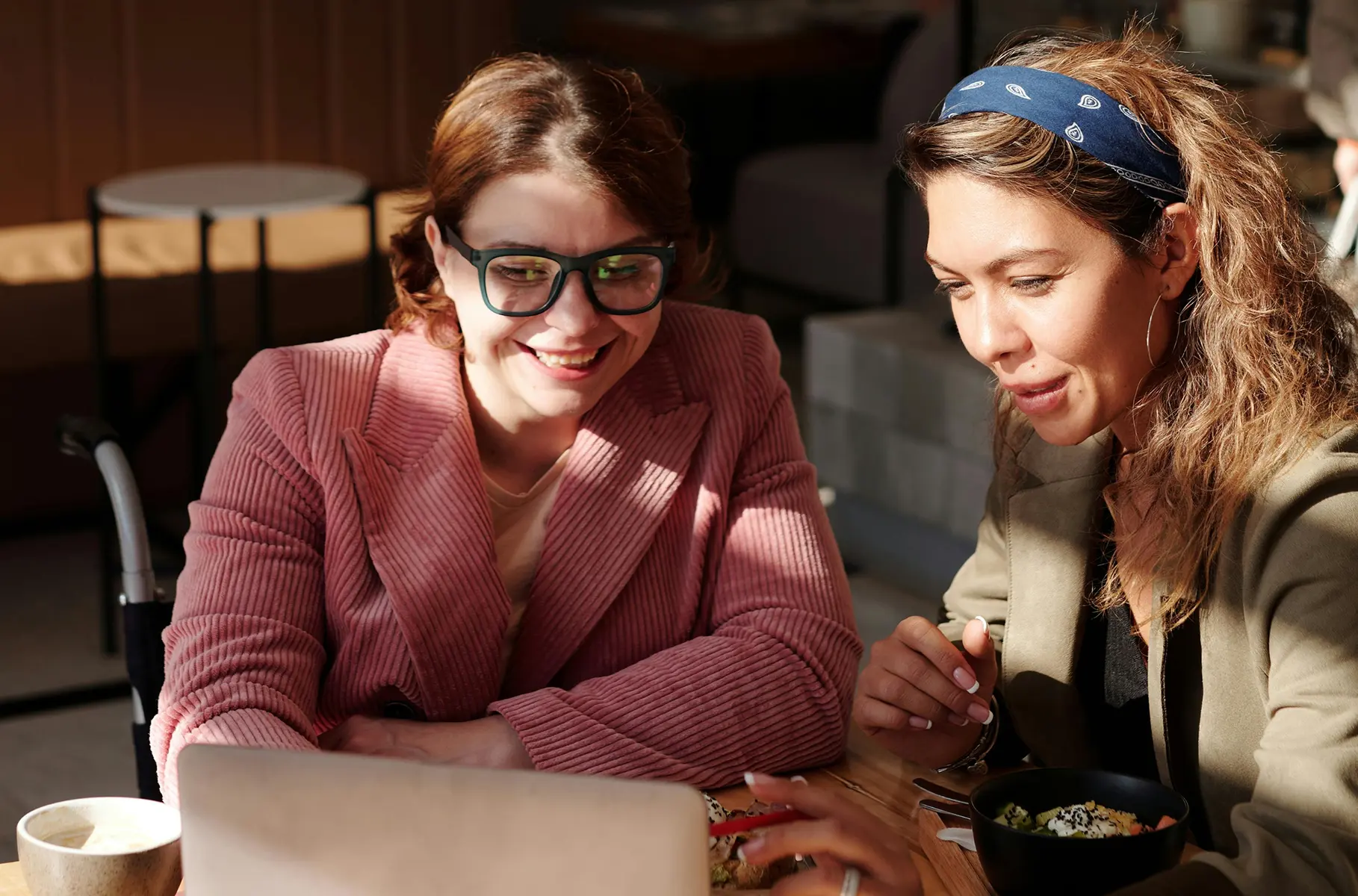 Two business women sitting at a café table, smiling and looking at a laptop together while discussing work over coffee and food.