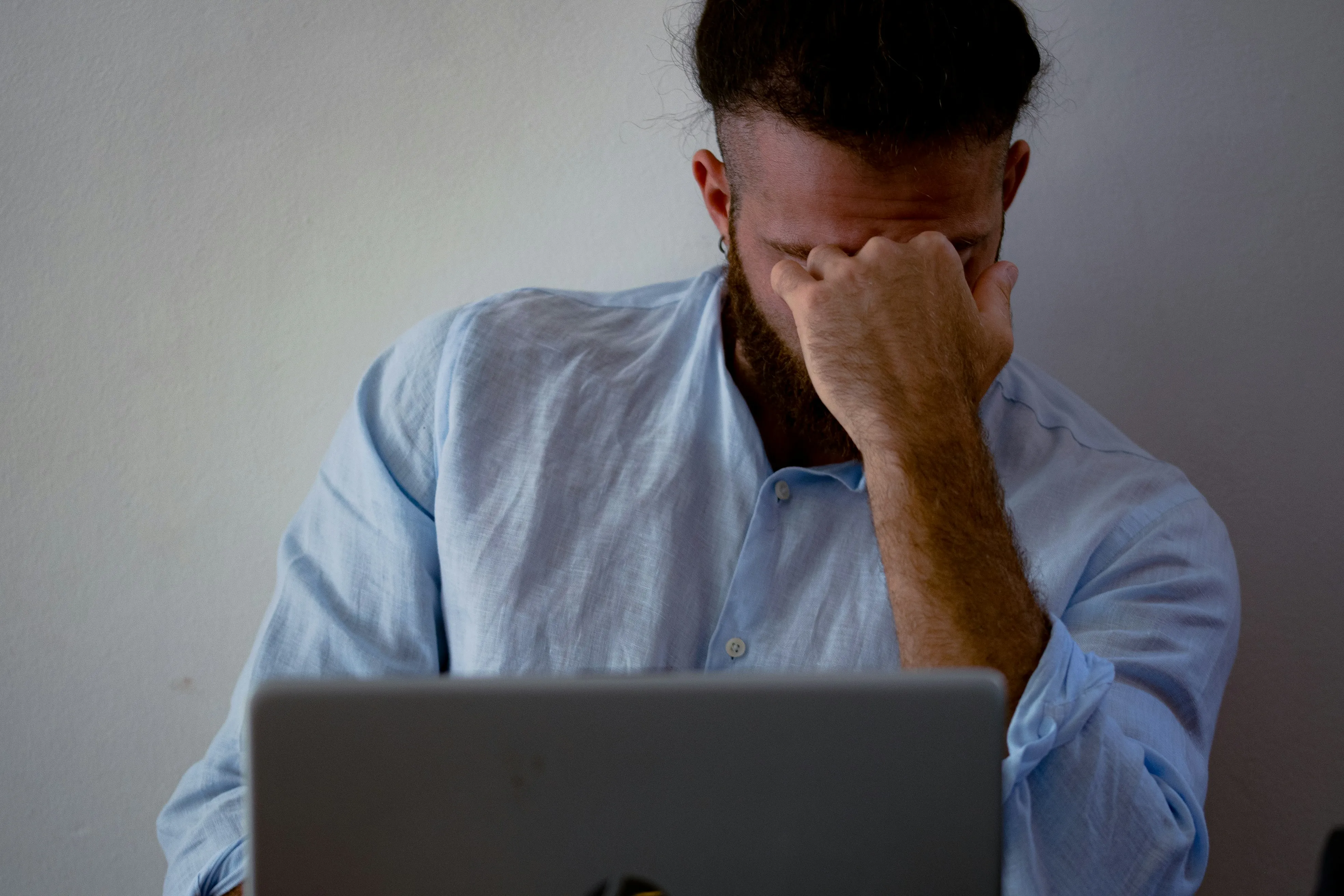 A person sitting with their head in their hand, appearing stressed, in front of a laptop on a desk, against a plain wall.