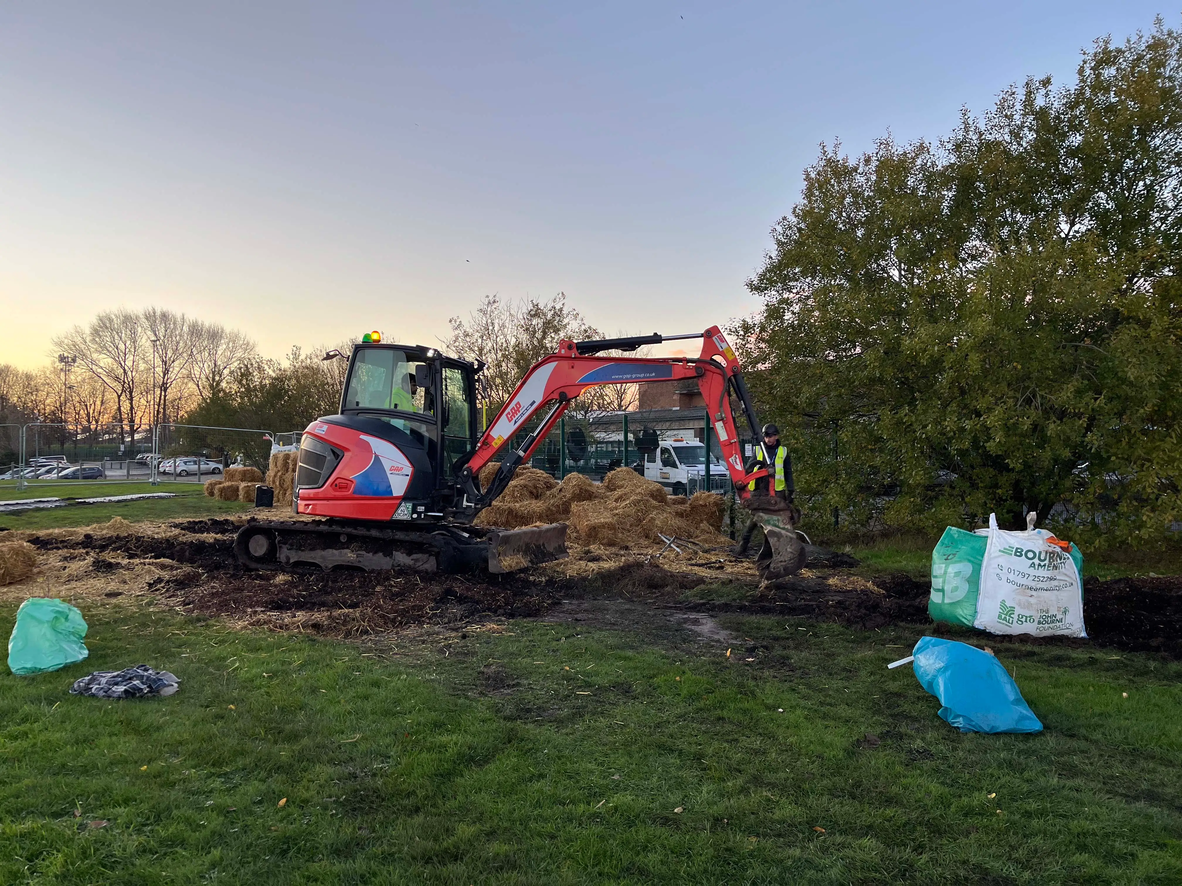 Excavator working in a field, surrounded by trees and equipment, during sunset with a colorful sky.