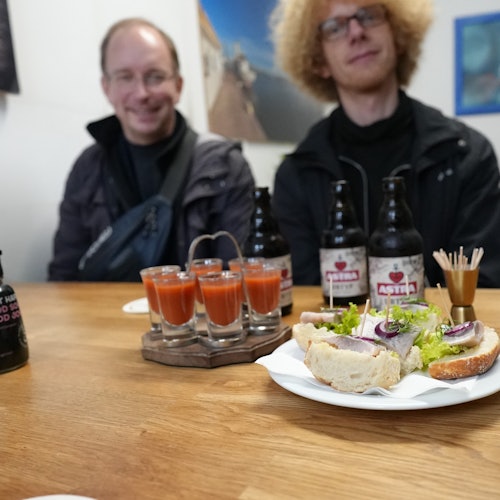 Two people sit at a table with sandwiches, tomato juice shots, and bottles of beer. Artwork on the walls in the background.