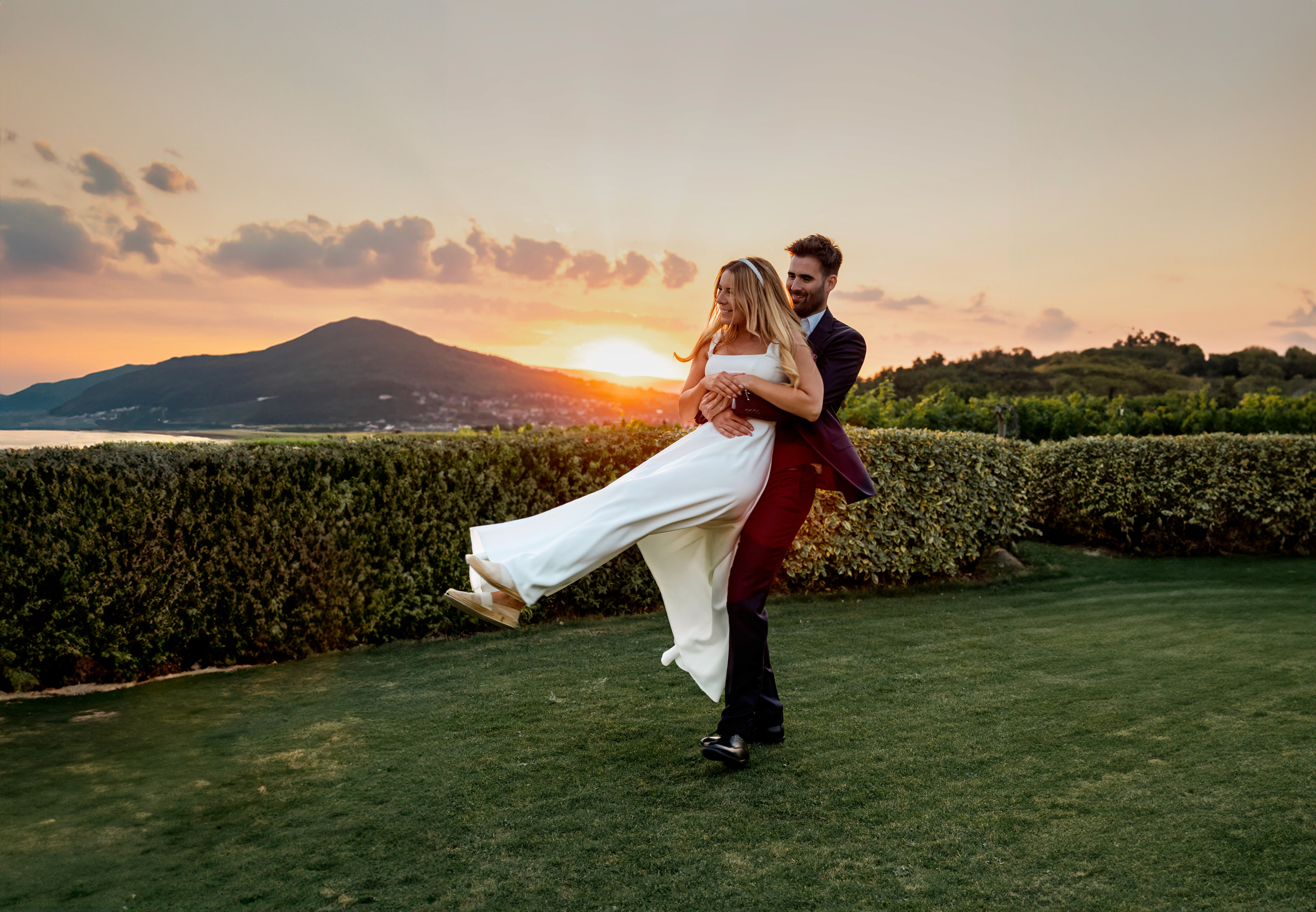 Vőlegény felemeli menyasszonyát, Balaton háttérrel és naplementével, vidám esküvői pillanatban.