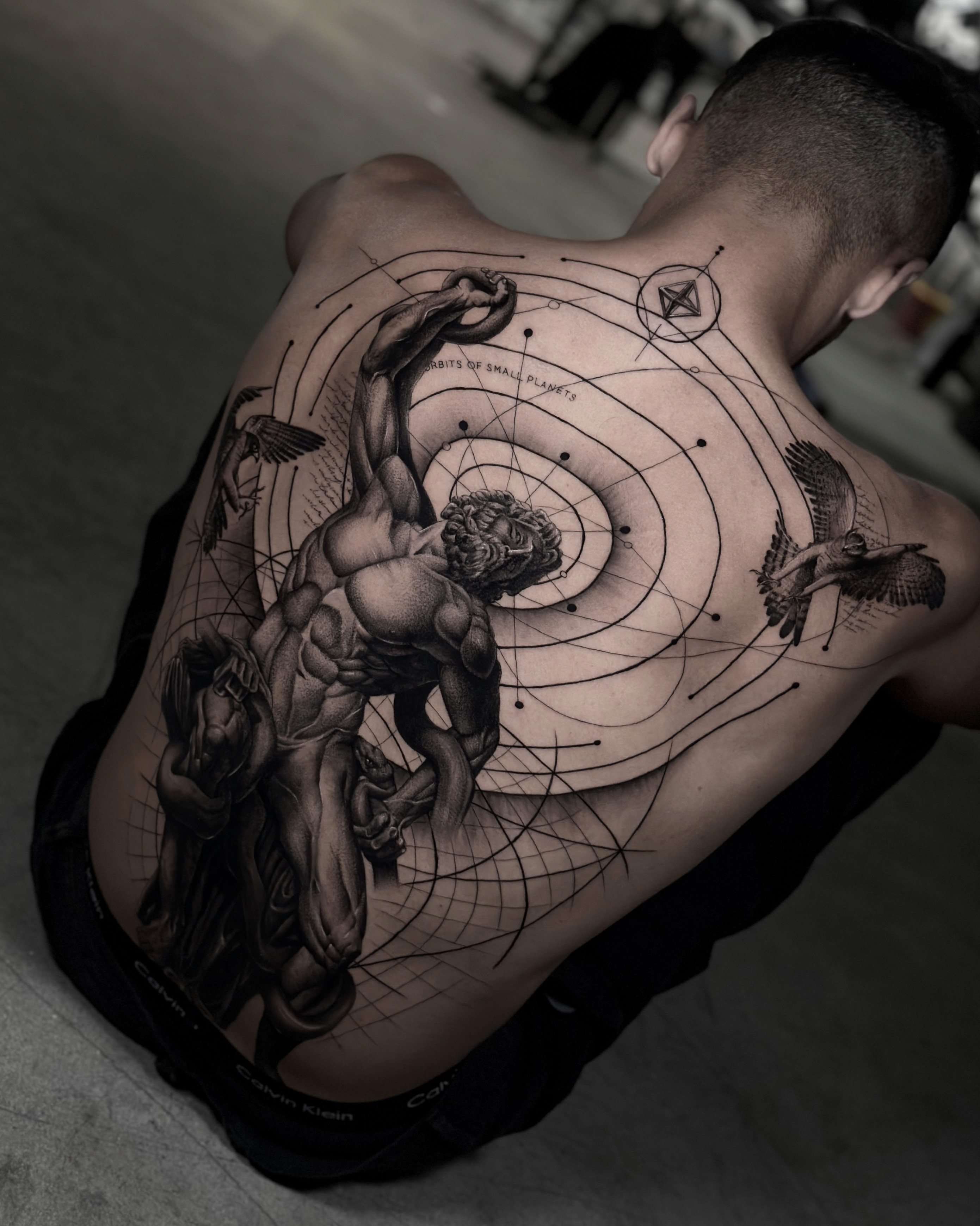 A detailed full-back tattoo of a dynamic, muscular male figure surrounded by geometric lines, astronomical elements, and flying birds, in a style inspired by classical sculpture and Renaissance art.