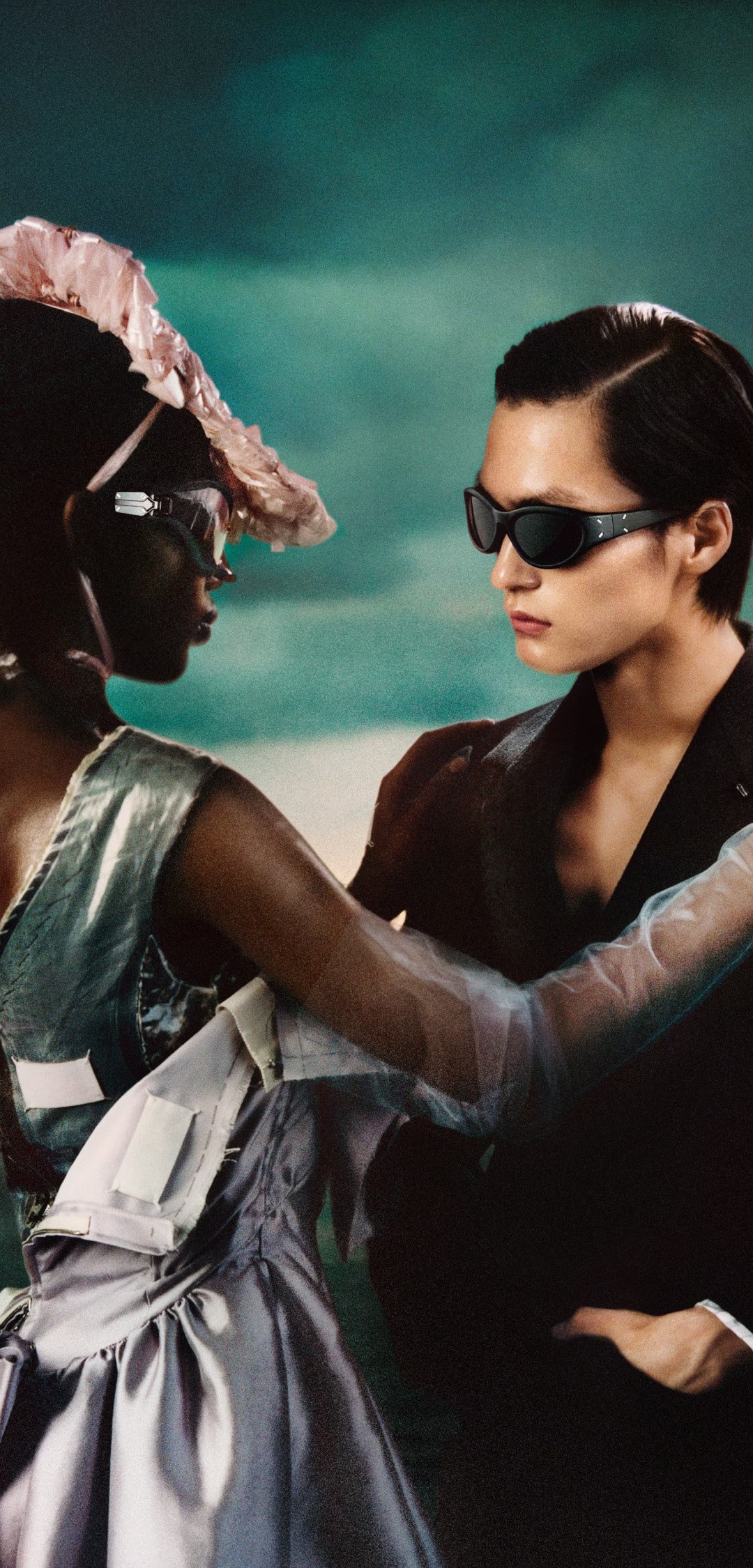 A fashionably dressed woman and man face each other closely, the woman wearing a dramatic hat and metallic dress, the man in dark sunglasses and a sleek outfit, set against a moody studio backdrop. Both are seen wearing Gentle Monster glasses.