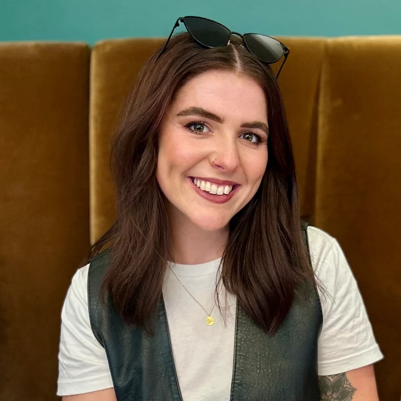 A person sits against a mustard‑coloured velvet bench wearing a white T‑shirt and dark vest. Sunglasses resting on their head and a teal wall behind create a relaxed, contemporary setting.