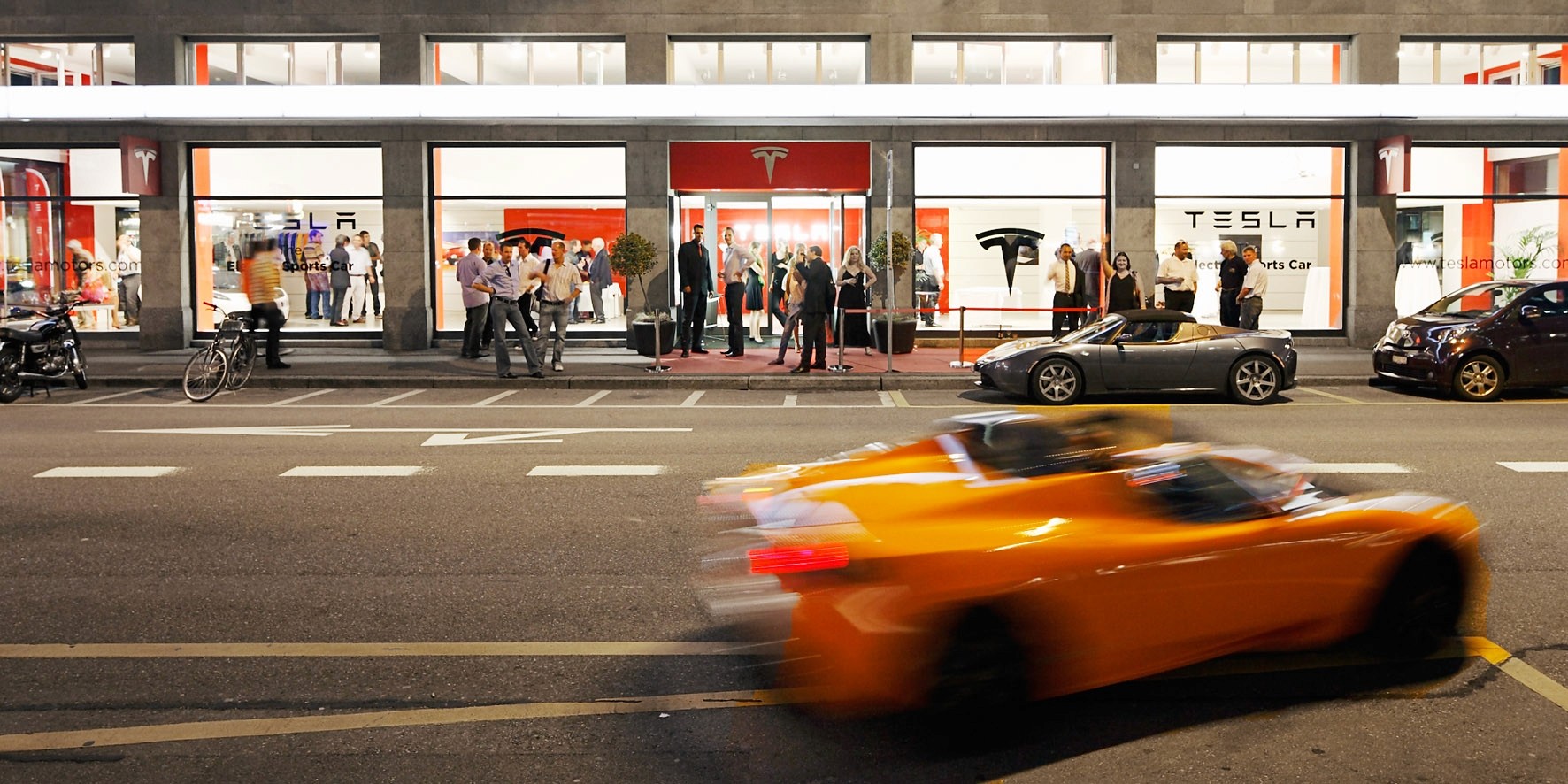 Exterior of a Tesla showroom with people gathered outside and an orange electric sports car driving by, representing the popularity of electric vehicles and Tesla's role in promoting electromobility in Switzerland