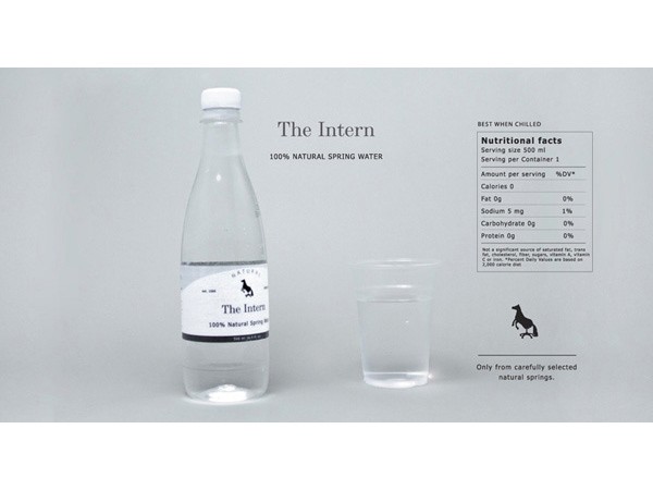 A clear plastic bottle labeled “The Intern” with 100% natural spring water sits beside a clear plastic cup filled with water. The bottle and cup are set against a light gray background. The label on the bottle includes the brand name “The Intern,” “100% Natural Spring Water,” and a horse silhouette. The Nutrition Facts panel is visible on the bottle, displaying details like serving size (500 ml), calories (0), fat (0), sodium (5 mg), carbohydrates (0g), and protein (0g). A small text box at the bottom reads “Only from carefully selected natural springs.”