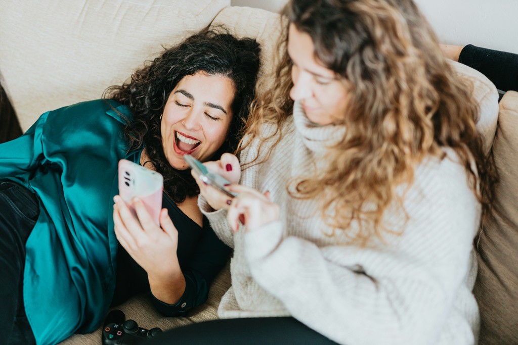 Two young women relax on a couch, smiling and engaging with their smartphones, exemplifying the joy and connection found through social media and digital communication.