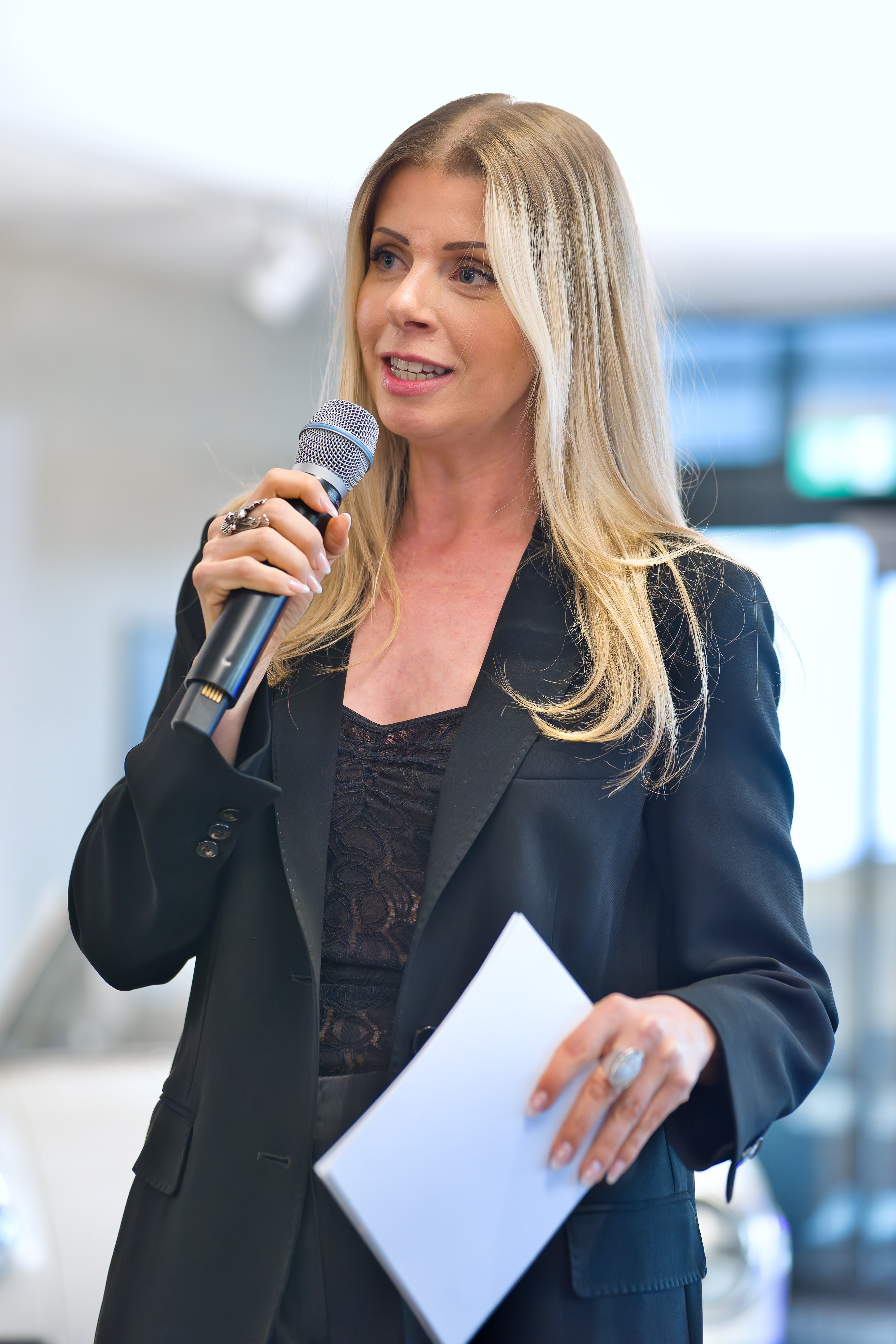 Blonde Moderatorin Désirée Lehmann, moderiert mit einem Mikrophon in der Hand und Moderationskarten in einem dunklen Anzug.