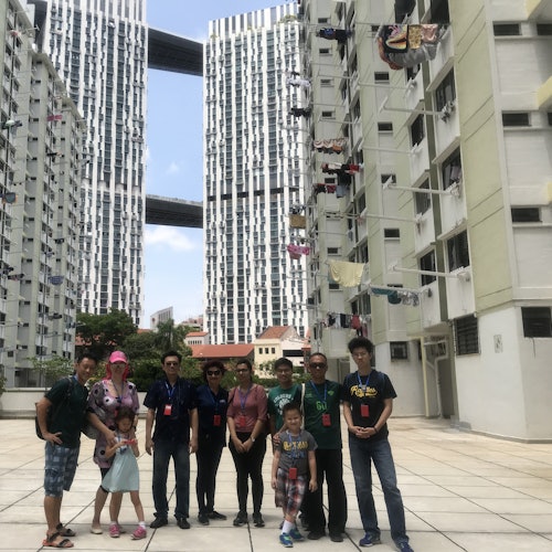 A group of people stands outdoors between two high-rise apartment buildings with sky bridges, with laundry hanging from balconies.