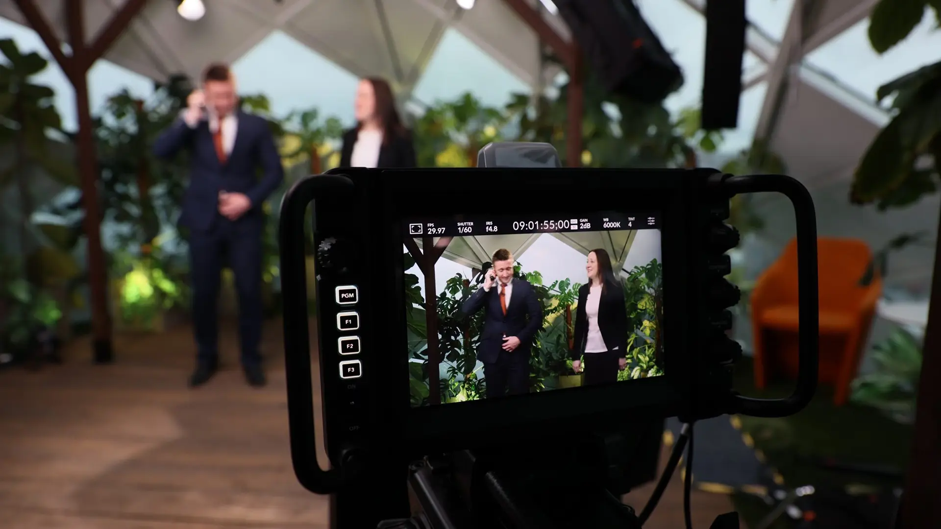 Camera monitor showing two presenters standing in a studio environment with plants and branded backdrop during a live event.