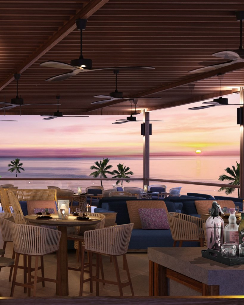 Interior of a modern restaurant or lounge with wooden ceiling and ceiling fans, featuring tables and chairs. The view shows a beach and palm trees at sunset with a colorful sky and ocean.