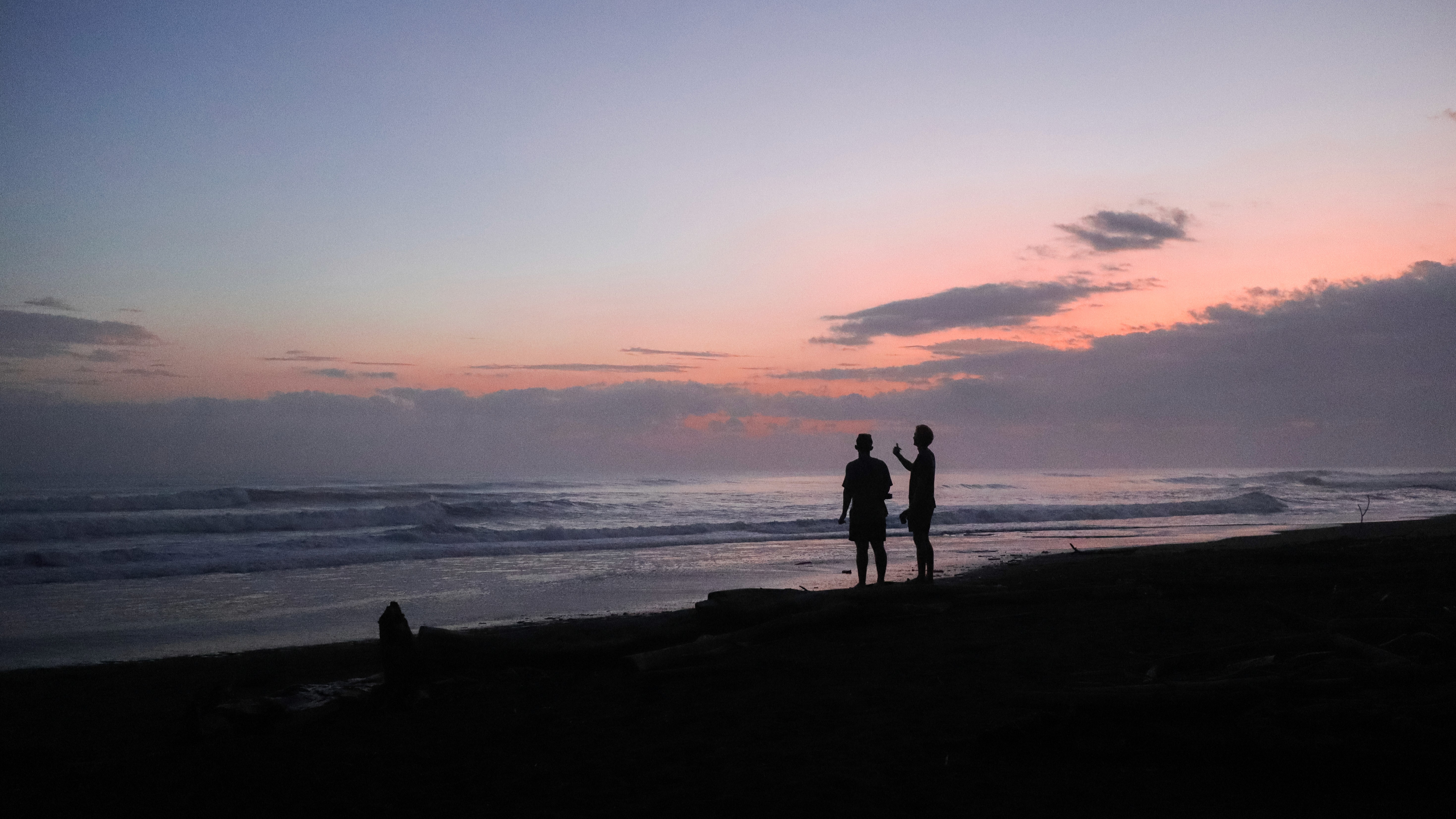 Two people standing on a beach at sunset, looking out toward the ocean.