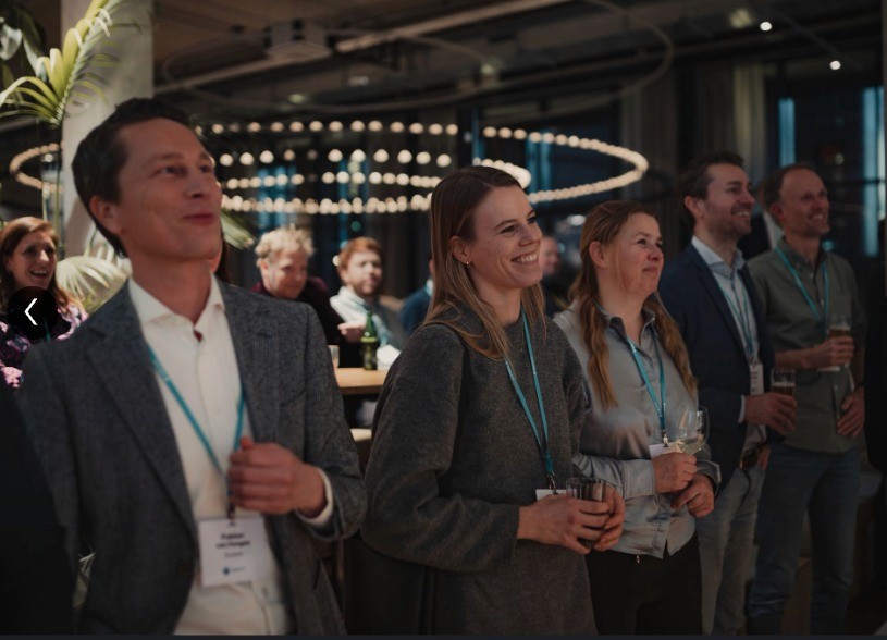 A group of people in business attire, listening and smiling at an event, with a lively background scene.
