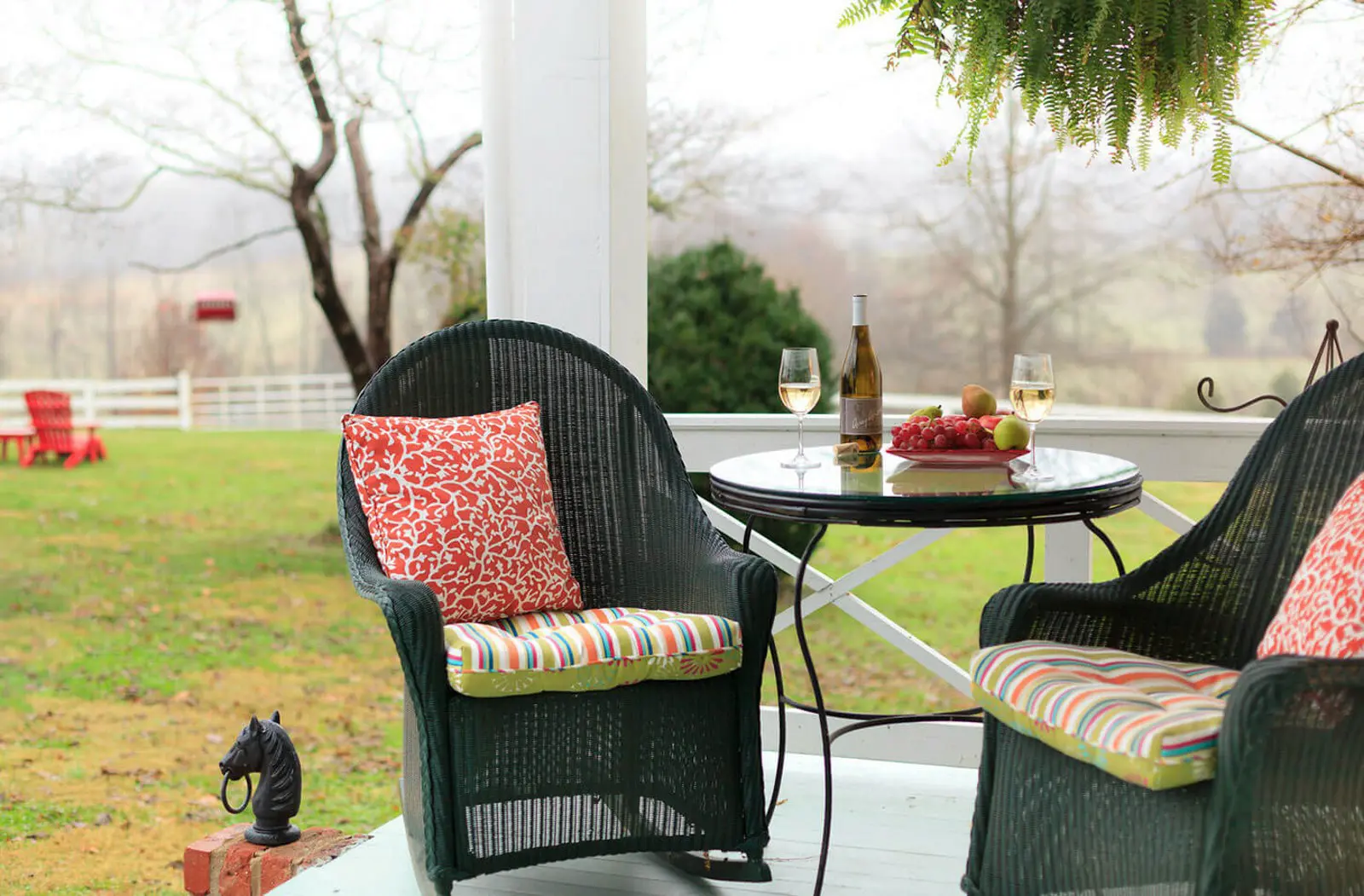 A cozy patio scene featuring two wicker chairs with colorful cushions, a small round table holding a bottle of wine and two glasses, surrounded by lush greenery and a scenic view of a rural landscape.