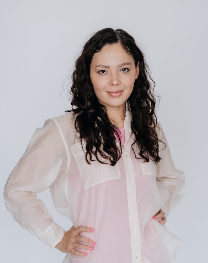 Kristina Lo wearing light pink and white attire with long, curly hair, standing upright against a plain background