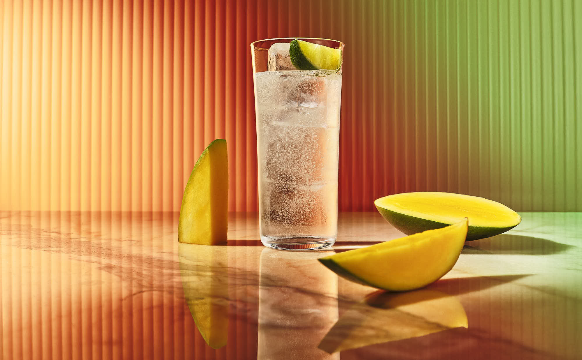 A refreshing drink in a tall glass with ice, garnished with lemon, beside fresh mango slices on a colorful background.