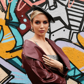 A blonde woman wearing a black beret and draped maroon jacket, posing in front of a colorful graffiti wall.