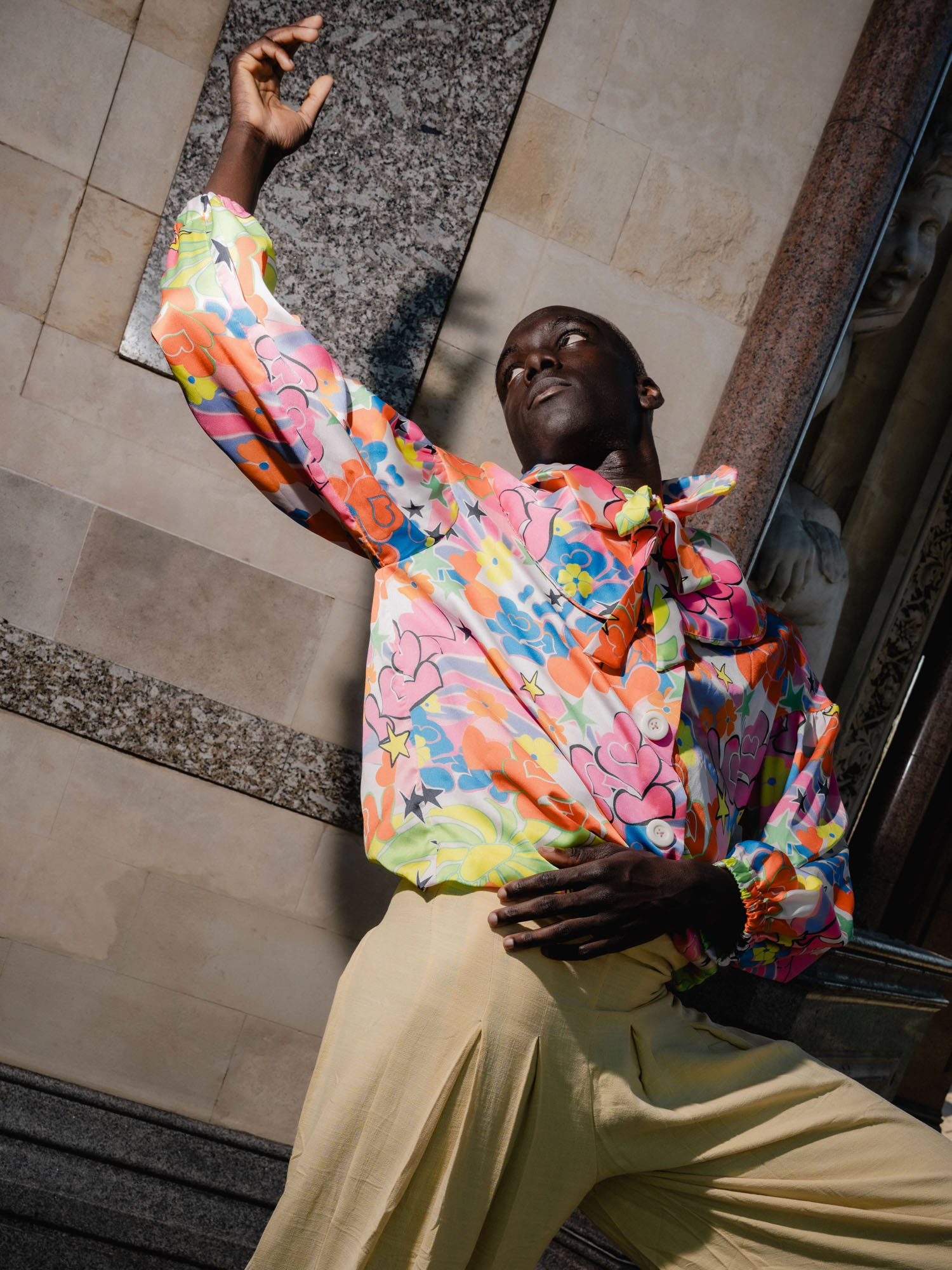 A person wearing a vibrant, floral-patterned shirt and cream-colored pants strikes a dynamic pose against a stone wall backdrop, exuding confidence and artistic expression.