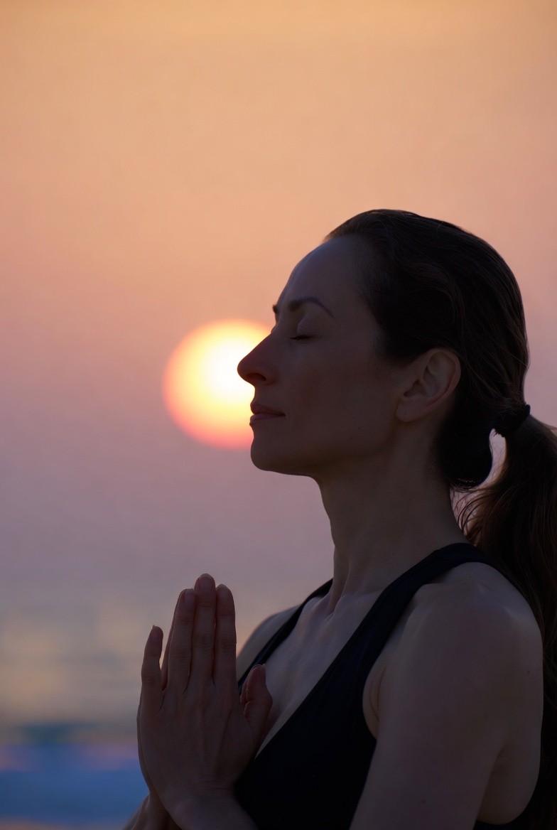 Woman meditating with eyes closed and hands in prayer position against a sunset sky