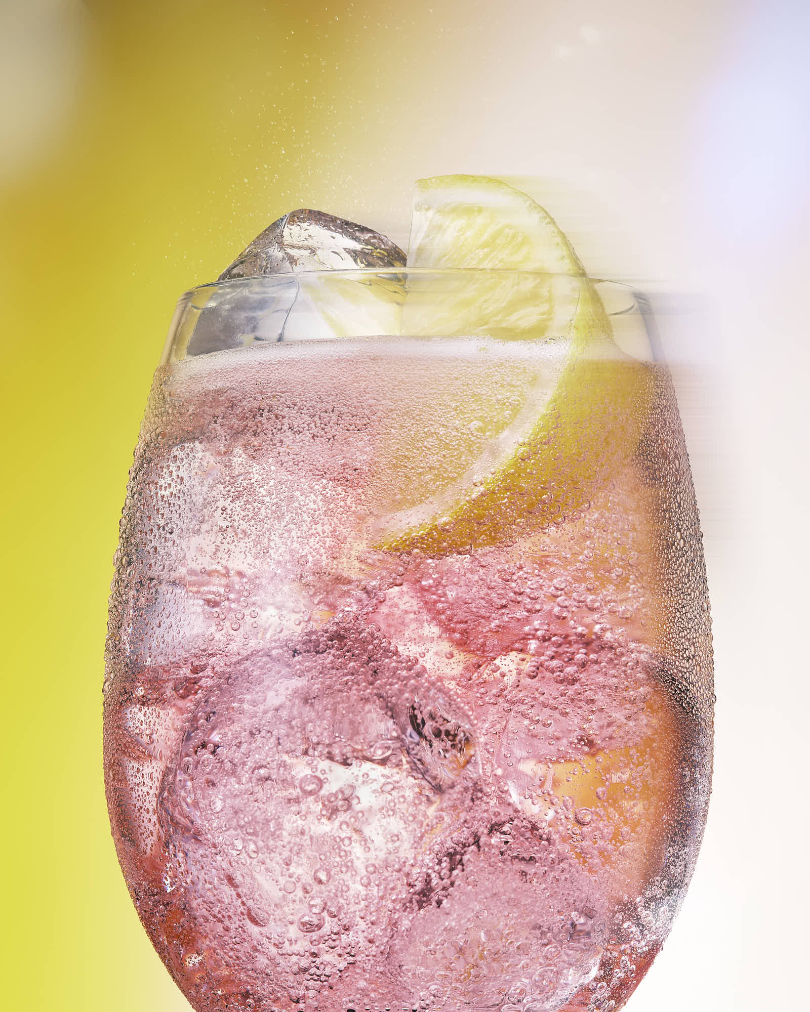 A refreshing pink cocktail with ice and a lemon slice, set against a soft yellow background.