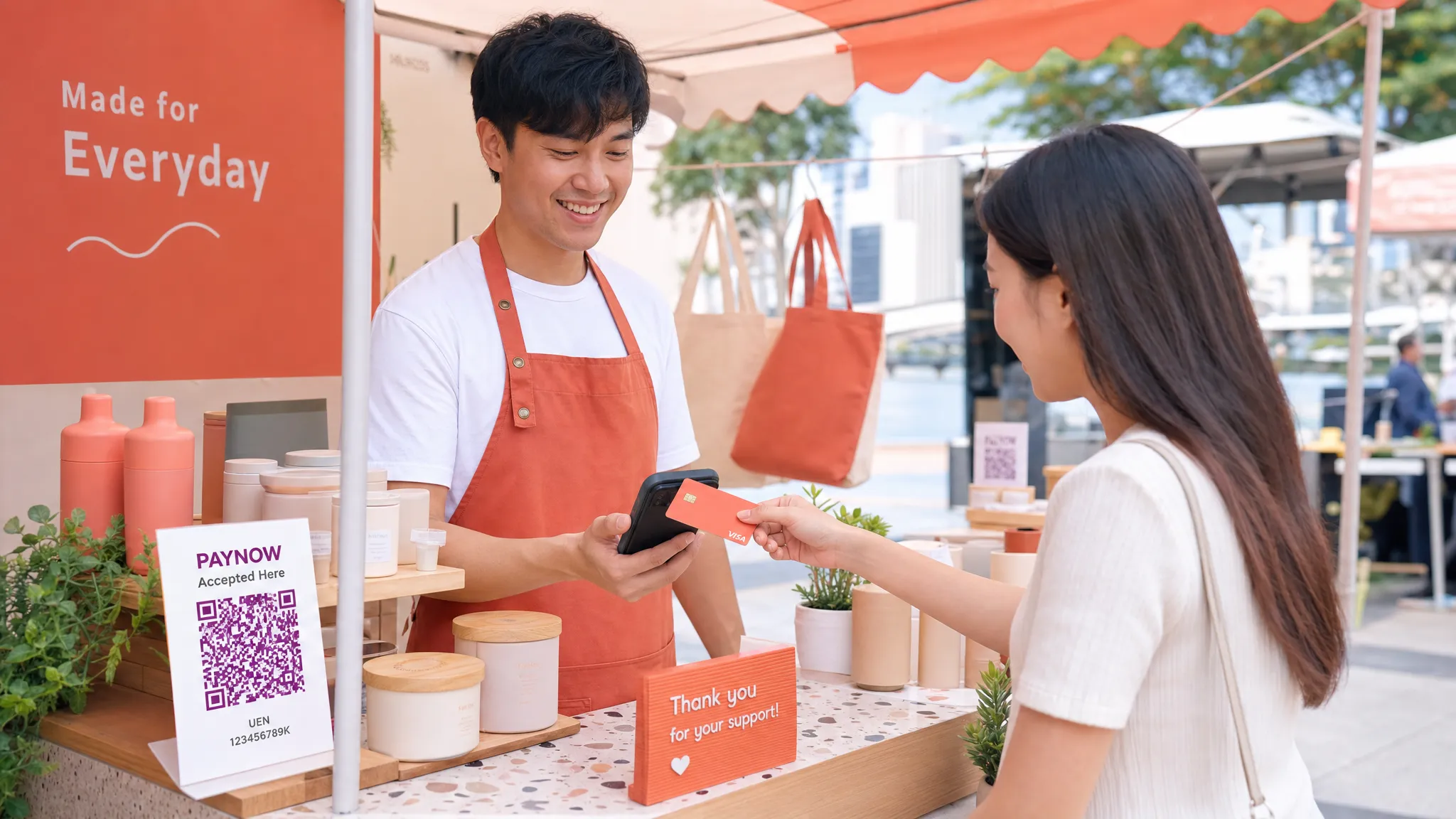 A Singapore pop-up merchant standing beside a compact market stall while a customer taps a contactless bank card on the back of the merchant’s smartphone to complete payment. The stall has neatly displayed products, a PayNow QR sign, and a clean checkout area.