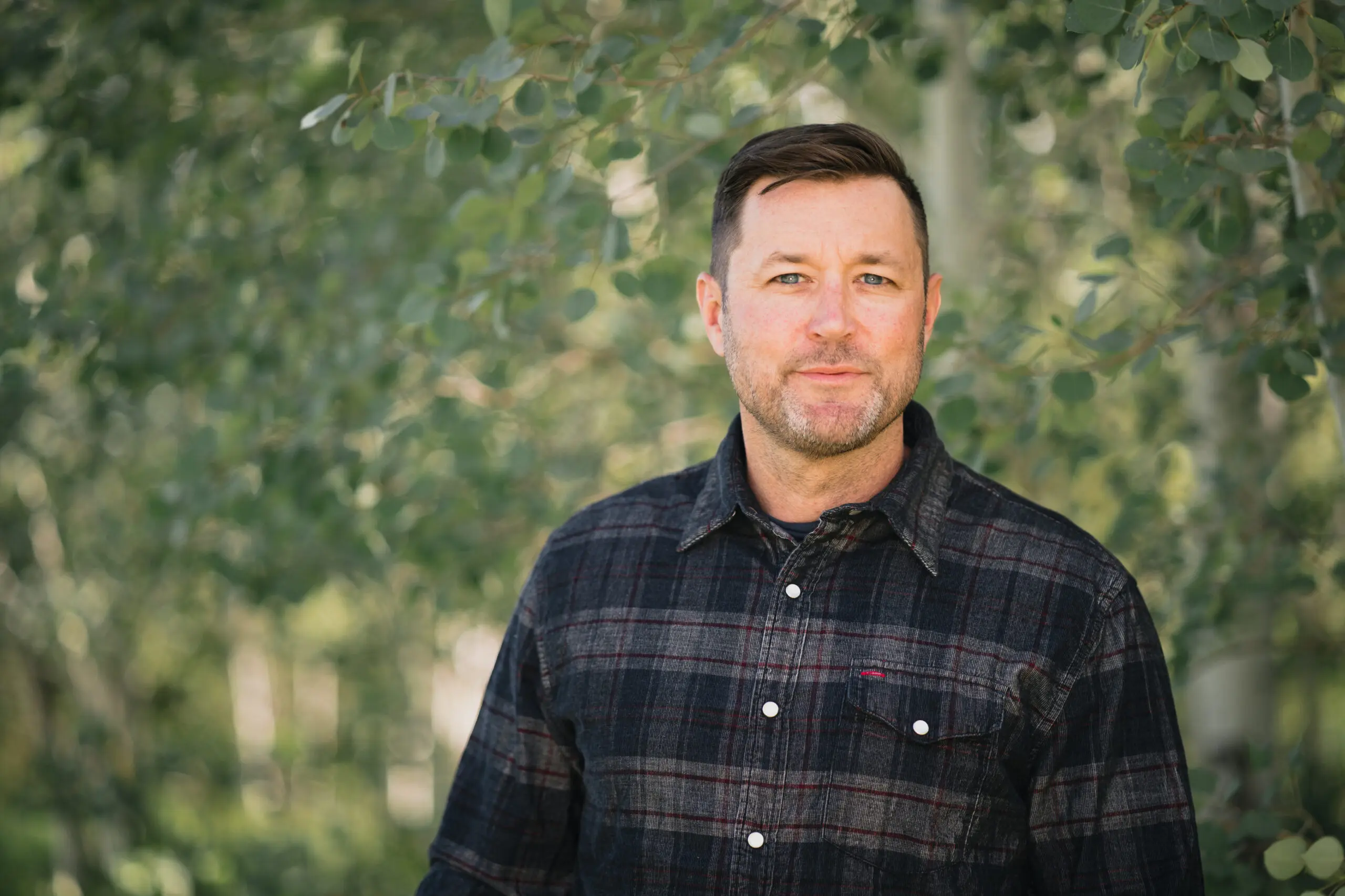 Man in a dark plaid shirt photographed against soft green foliage.