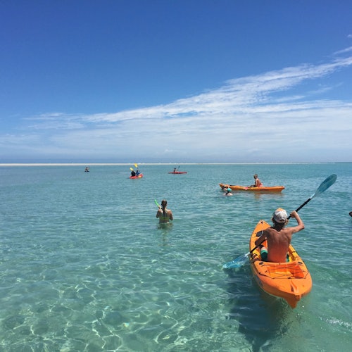 People kayaking and swimming in clear, shallow ocean water under a bright blue sky with scattered clouds.