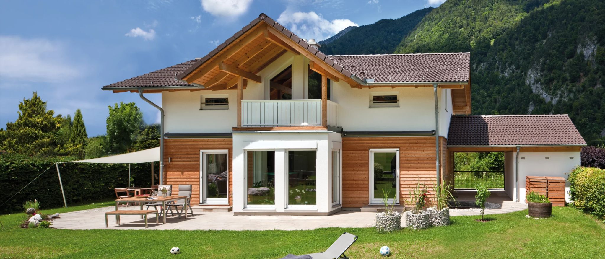 Modernes Einfamilienhaus mit Holzfassade, großzügiger Terrasse und Gartenanlage, umgeben von Berglandschaft.