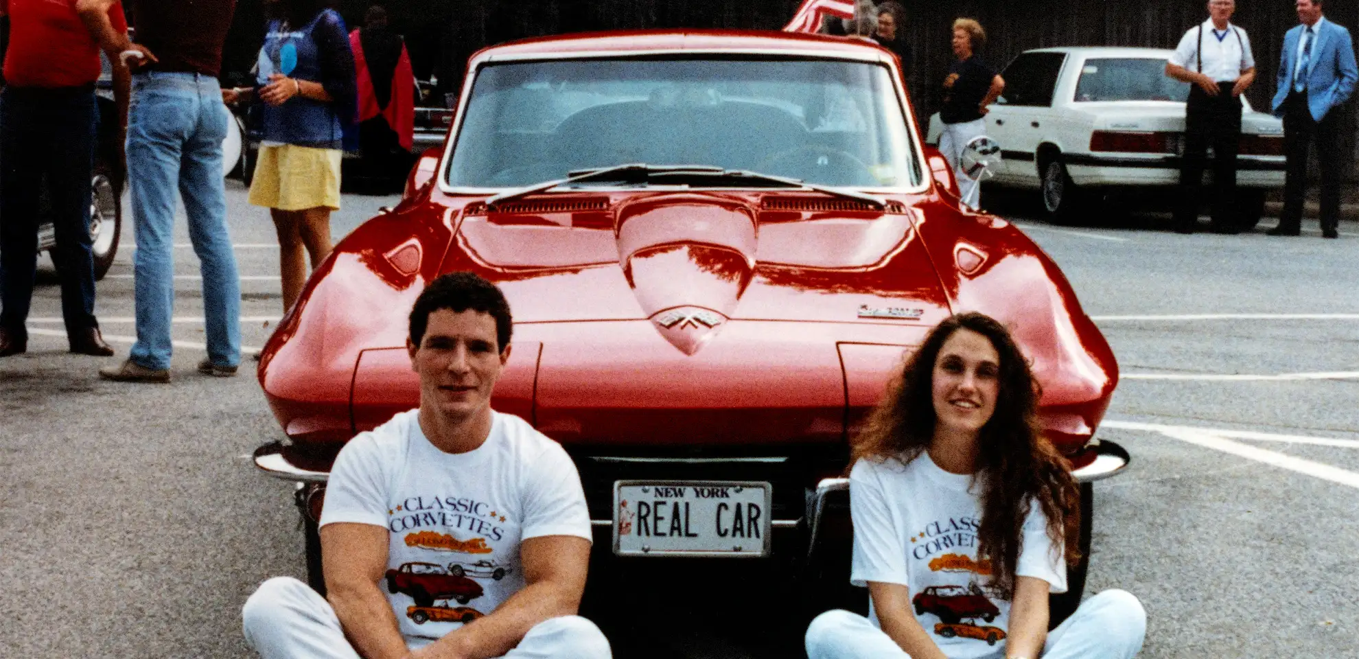 A man and woman sit by a red Corvette with a “REAL CAR” plate, wearing “Classic Corvettes” T-shirts at a car show.